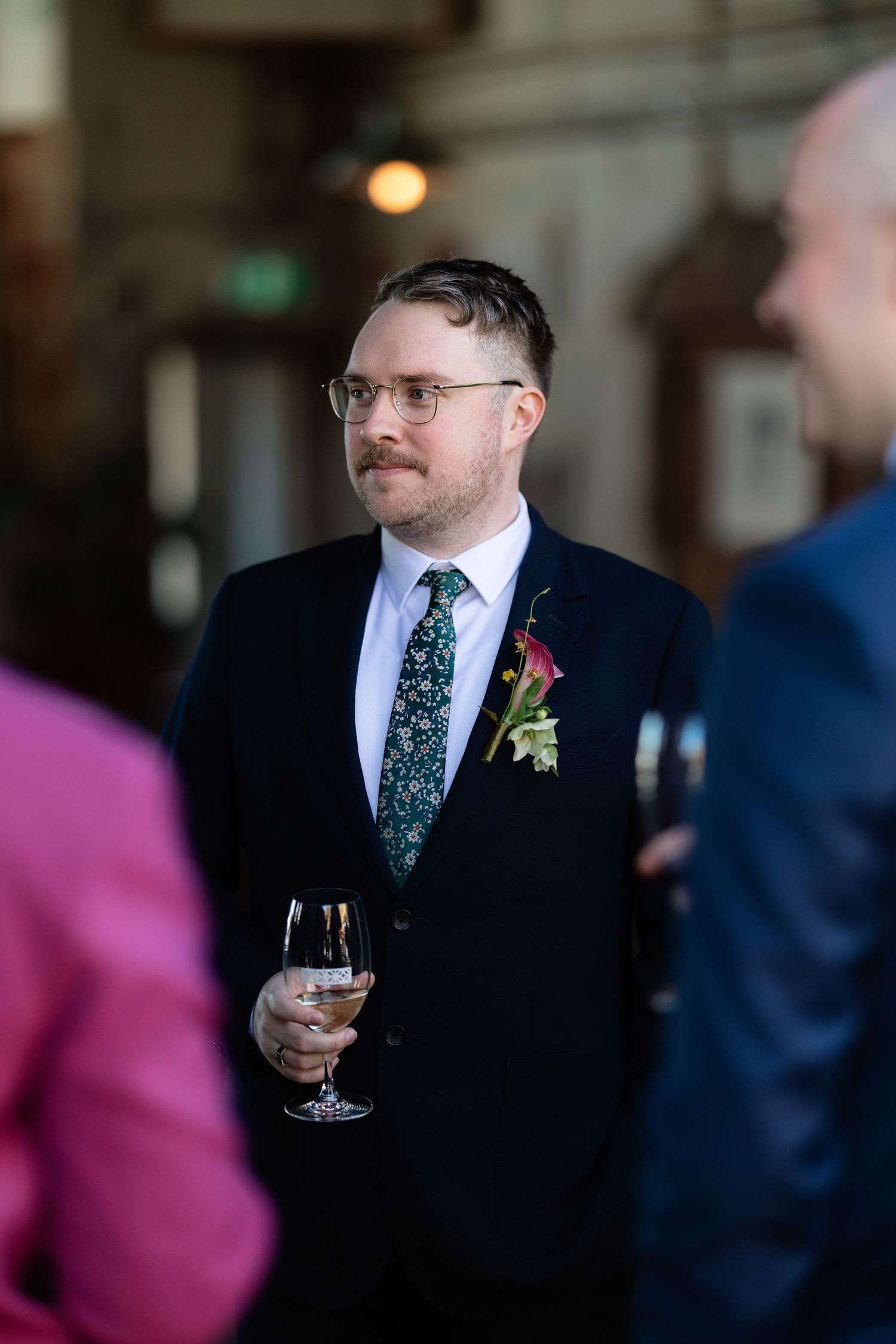 Groom in navy suit with floral tie and pink calla lily boutonniere holds champagne glass at wedding reception.