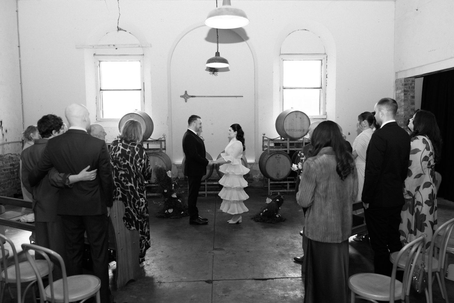 Black and white photo of intimate winery wedding ceremony with small guest gathering among wooden barrels.