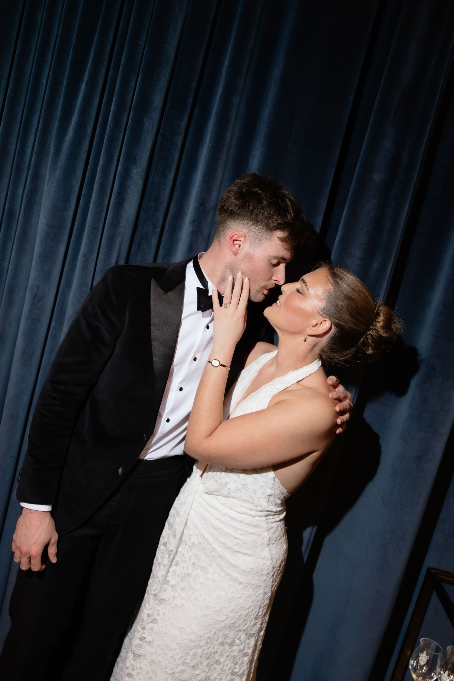 Bride and groom sharing a romantic kiss in front of blue velvet curtains, wearing wedding dress and tuxedo.