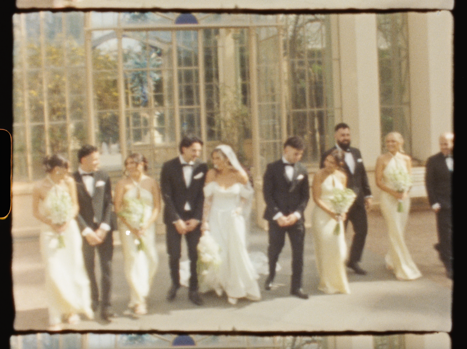 Wedding party poses outside elegant glass conservatory, bride in white gown with groom and bridesmaids in gold dresses.