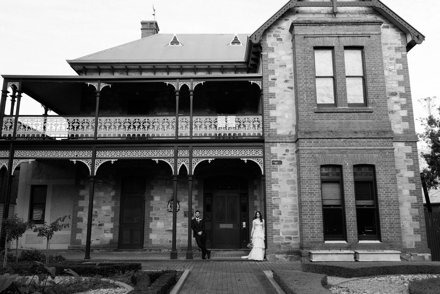 Bride and groom posing at entrance of historic stone Victorian mansion with ornate iron balcony in black and white.