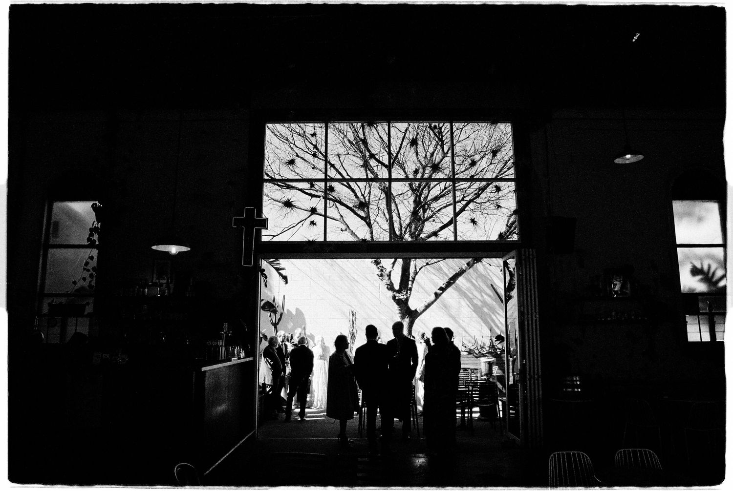 Black and white silhouette of people gathered in a doorway with a bare tree and bright light behind them.