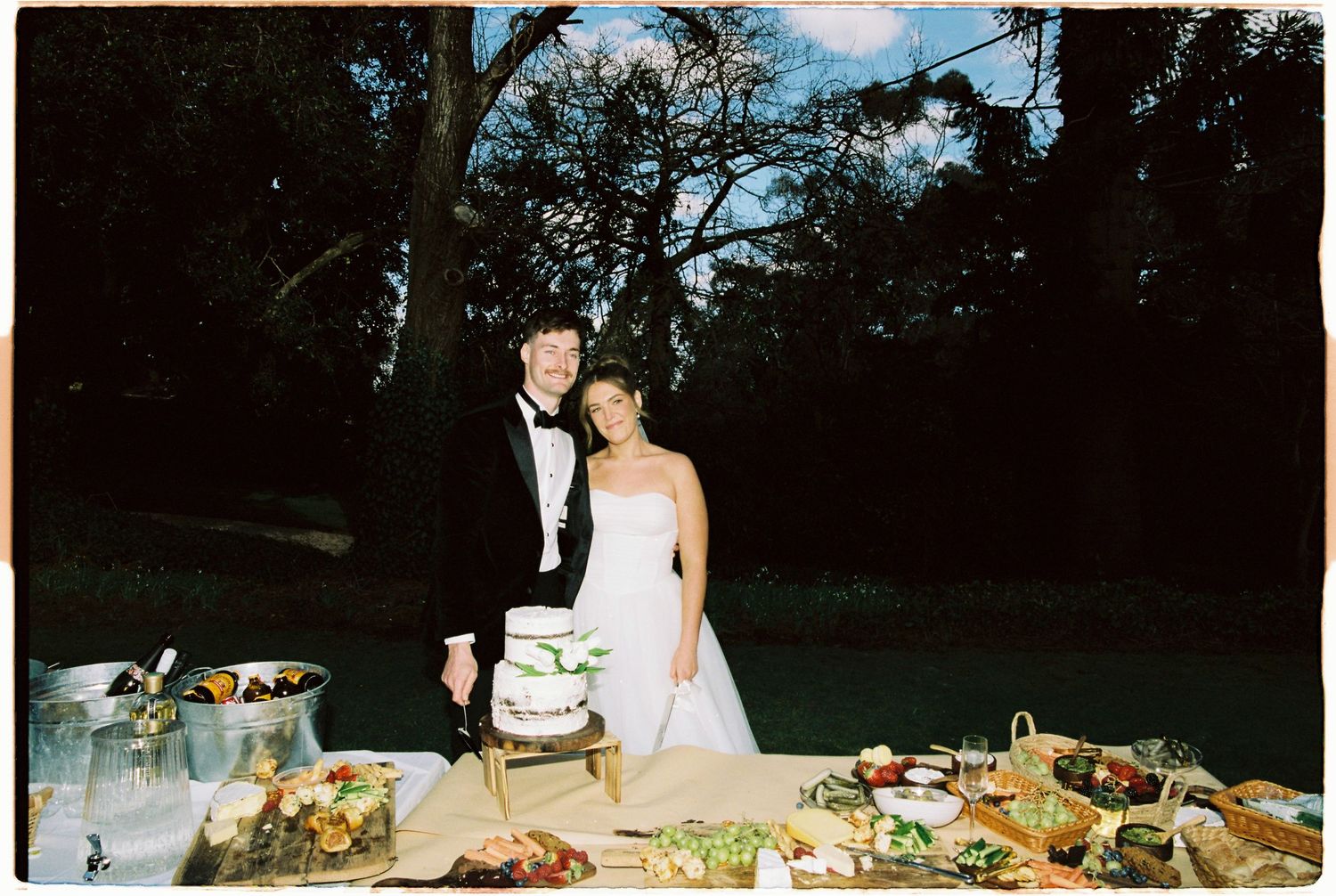 Bride and groom posing together at outdoor wedding reception table with cake and floral arrangements.