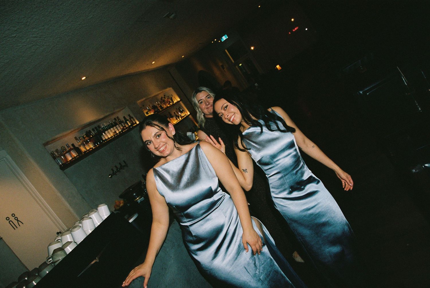 Three bridesmaids in matching silver satin dresses posing together near the wedding bar.