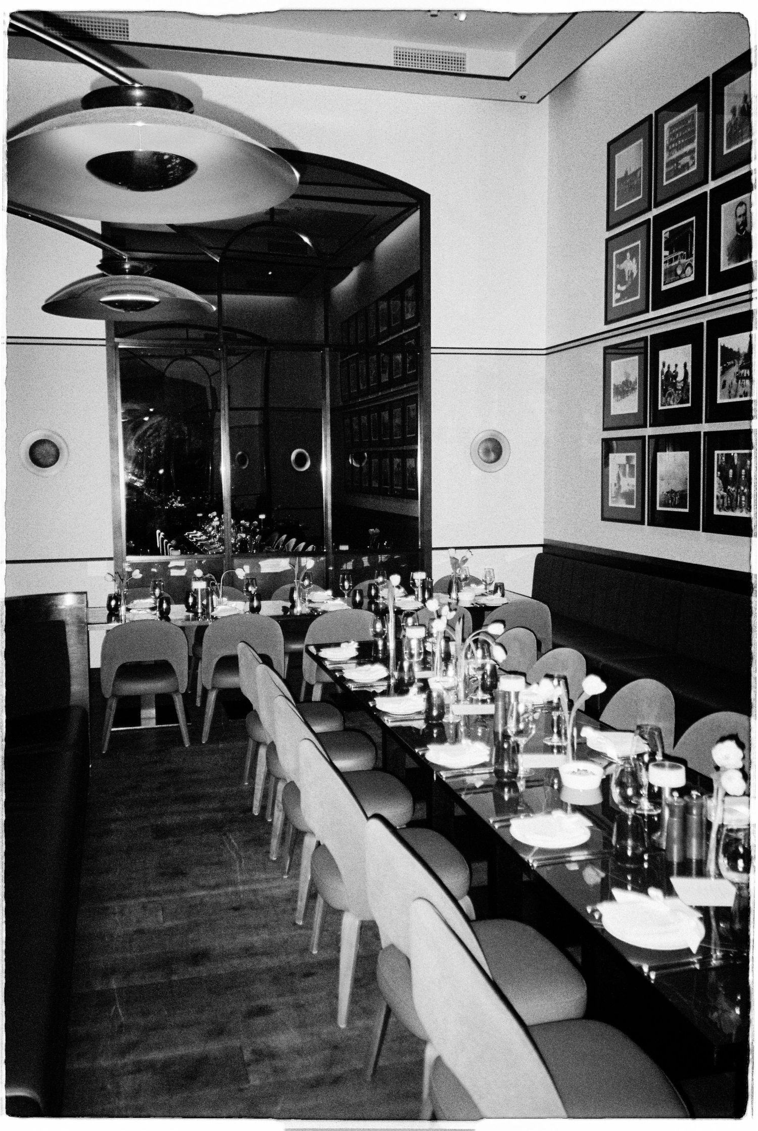 Black and white photo of an elegant restaurant interior with long dining tables, framed artwork on walls, and pendant lighting.
