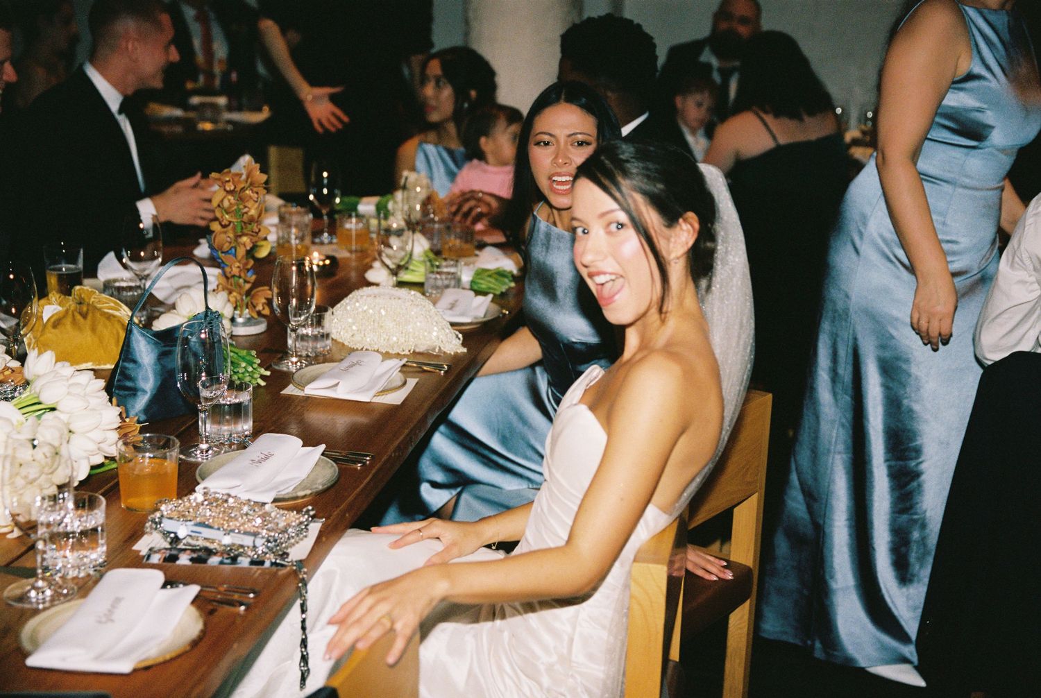 Elegant wedding reception dinner with guests in satin gowns seated at a decorated table with floral centerpieces.