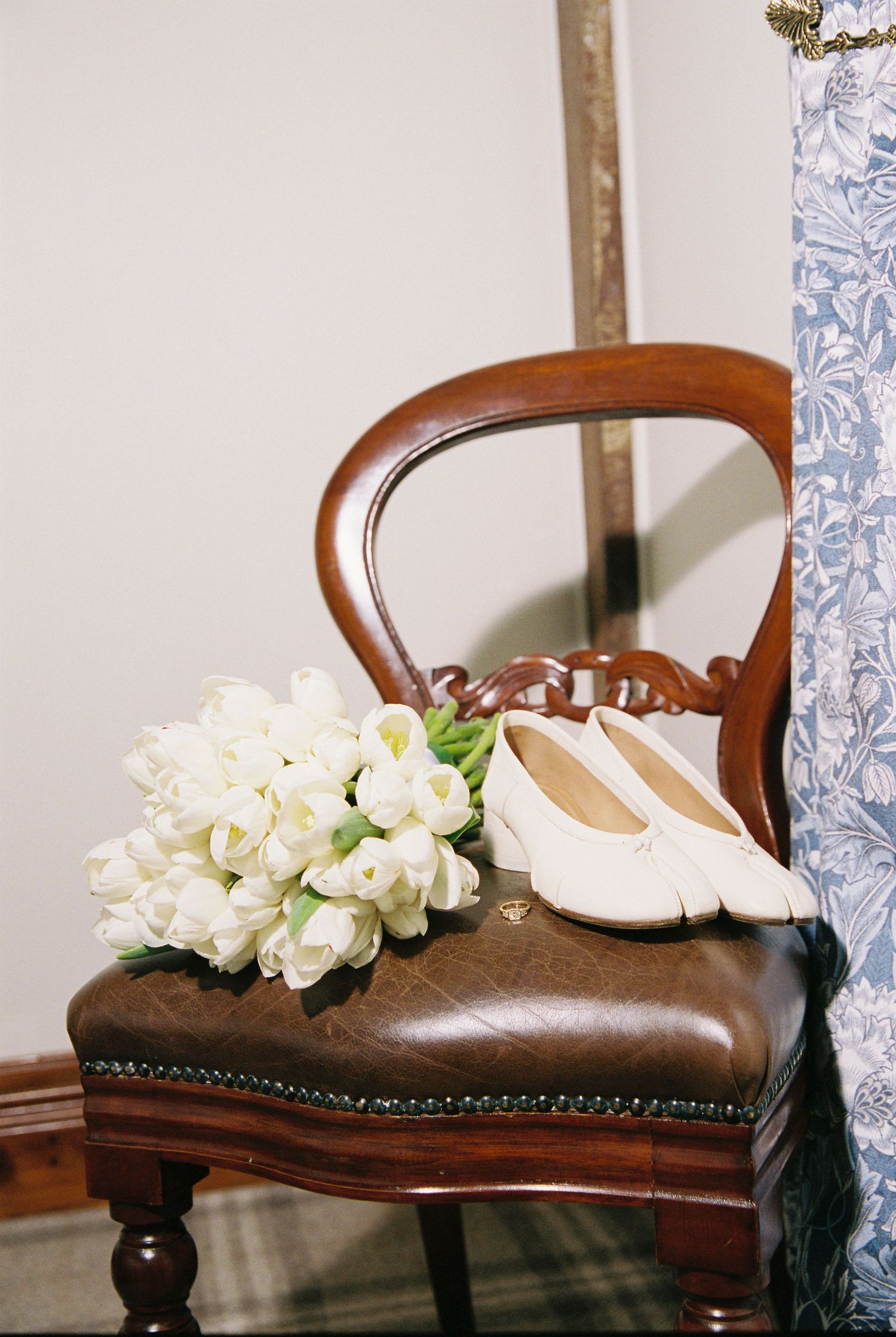 White tulip bridal bouquet and ivory flat shoes resting on a vintage leather chair.