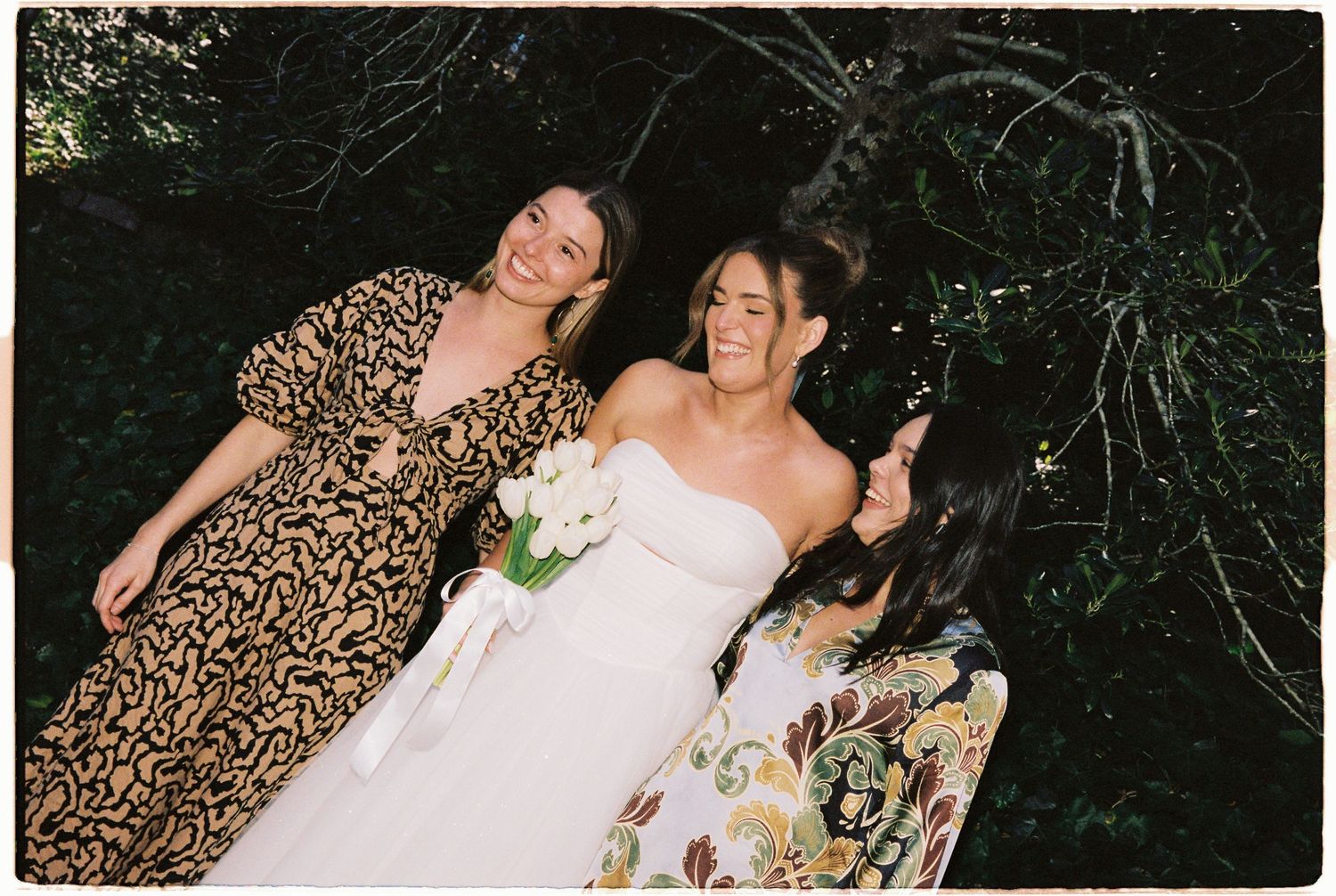 Bride in white strapless gown poses with two bridesmaids in floral dresses holding bouquet.