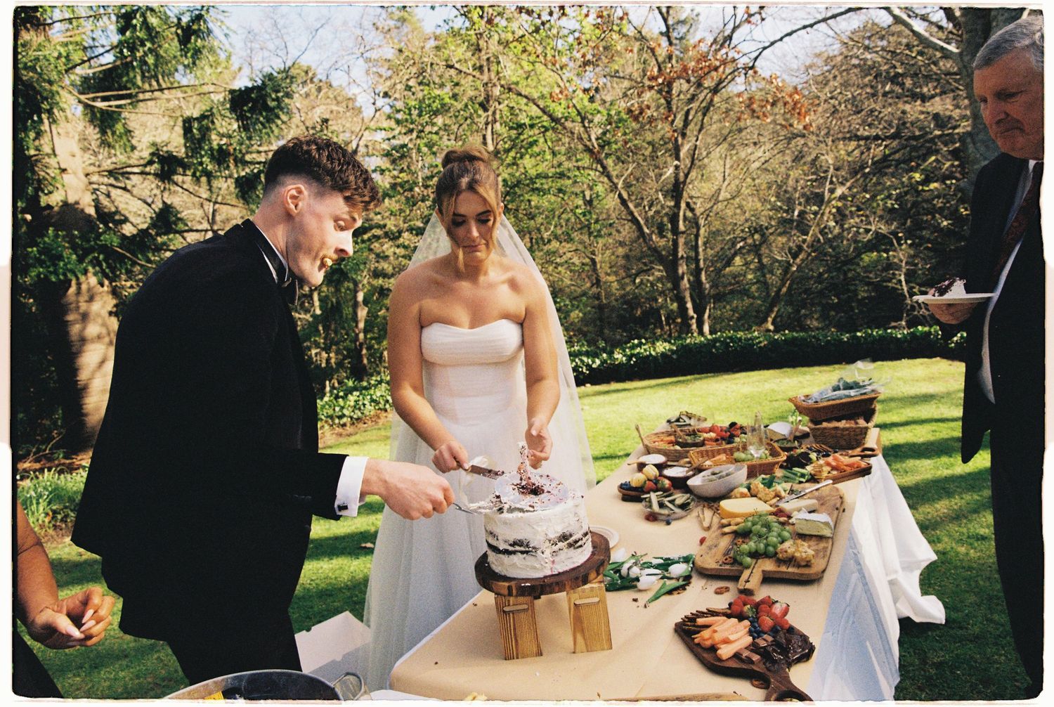 Bride and groom cutting wedding cake outdoors at garden reception table with food spread.