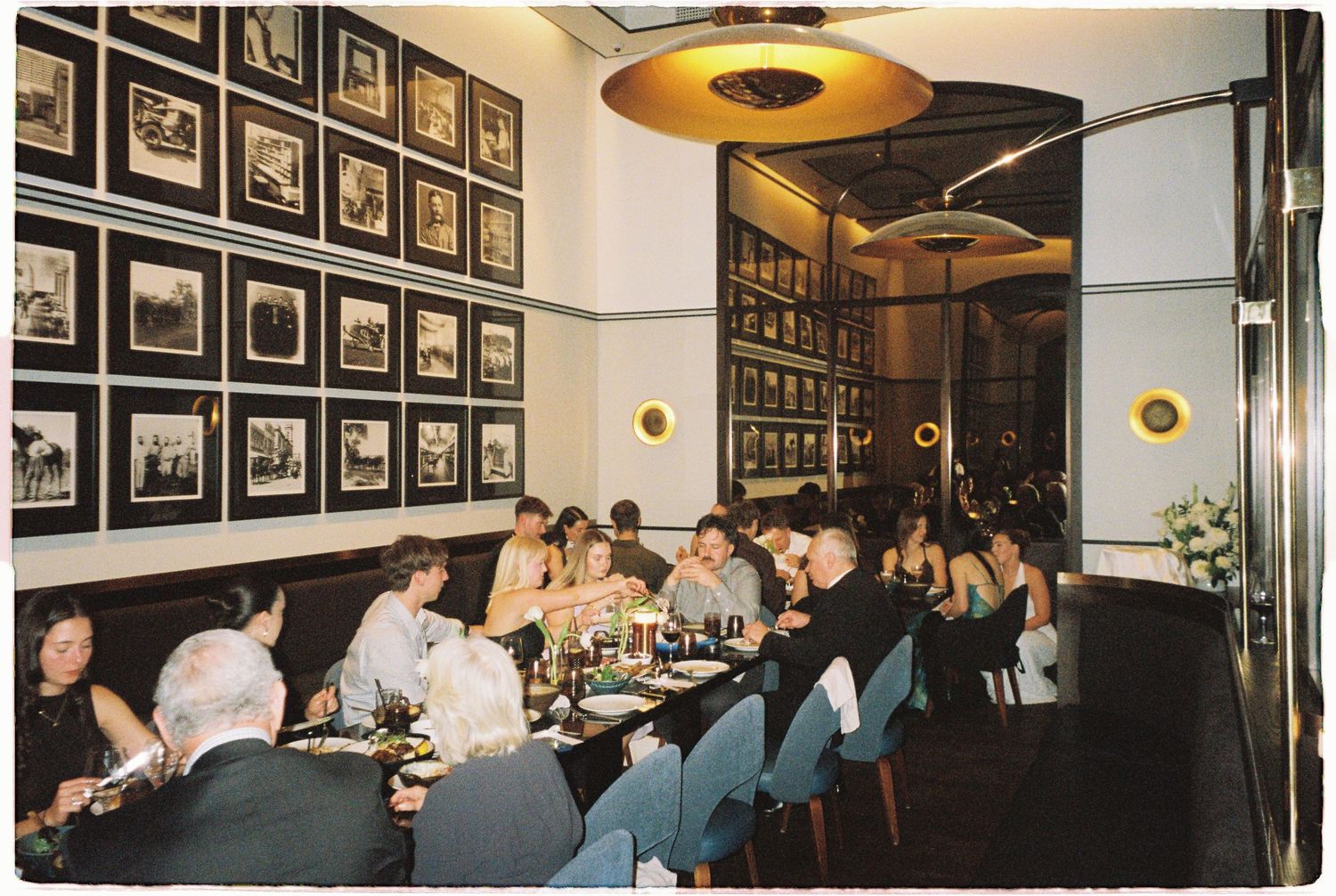 Wedding reception guests dining at a long table in an elegant restaurant with framed photos on walls.