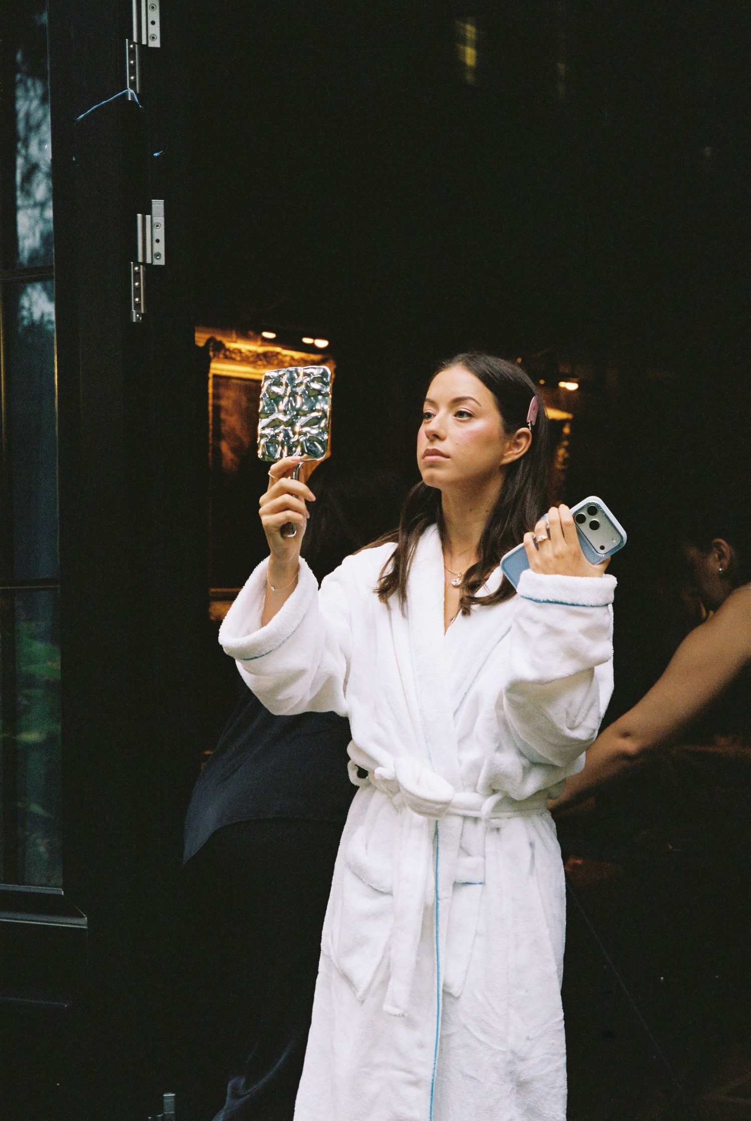 Woman in white robe holding phone and compact mirror outside at night.