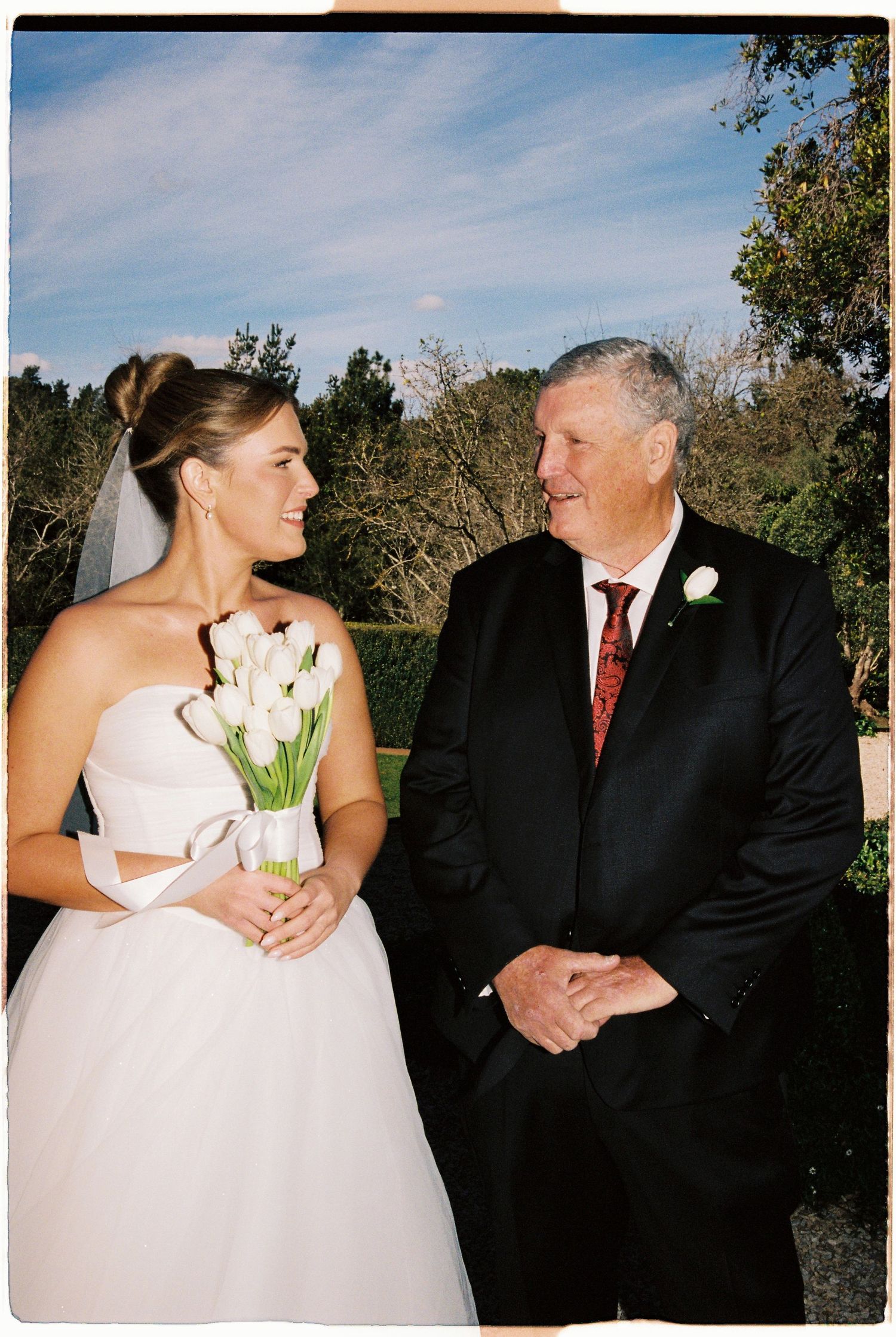 Bride in white strapless gown holding tulip bouquet smiles at older man in black suit with boutonniere at outdoor wedding.