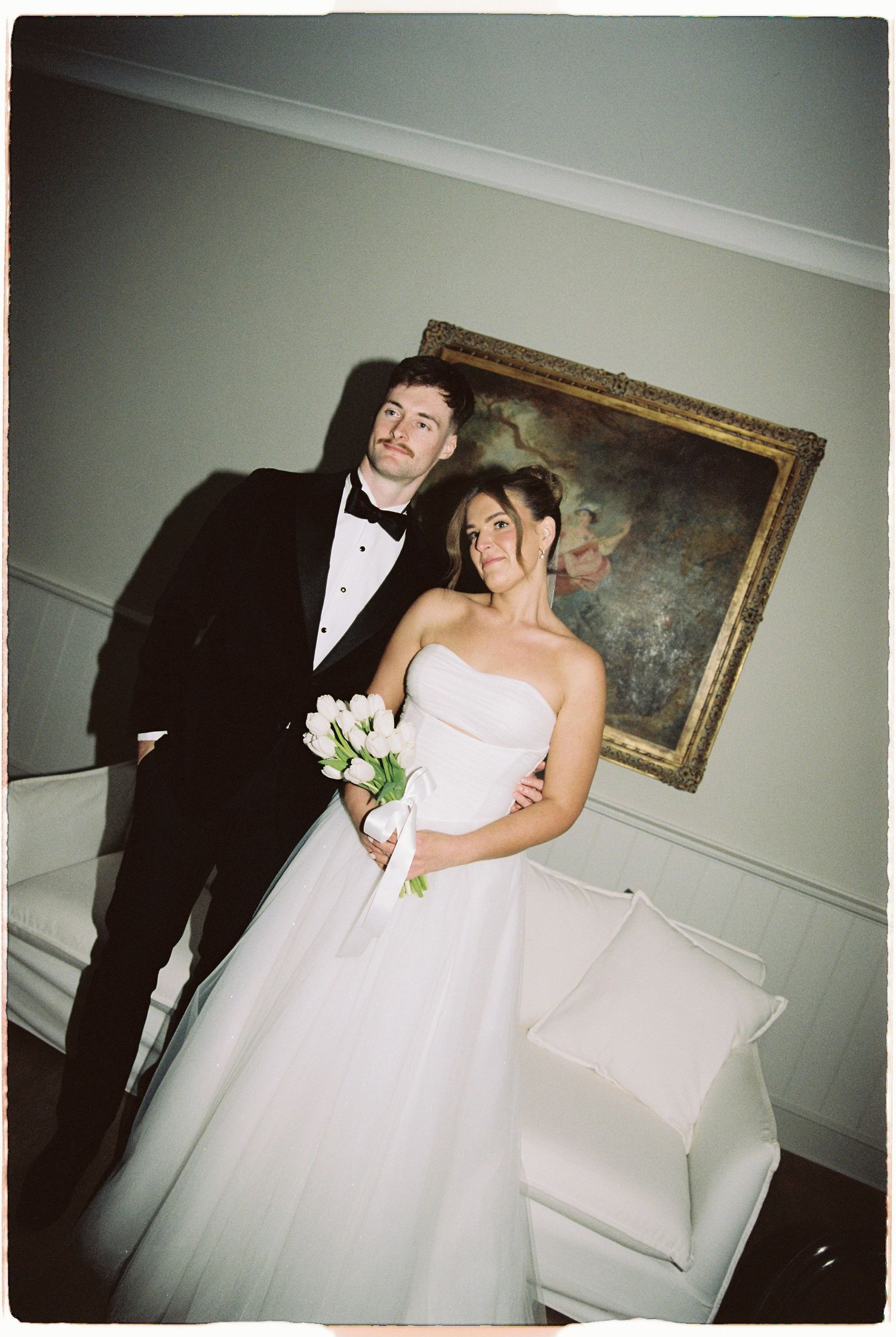 Bride and groom in formal wedding attire pose indoors before classic oil painting.