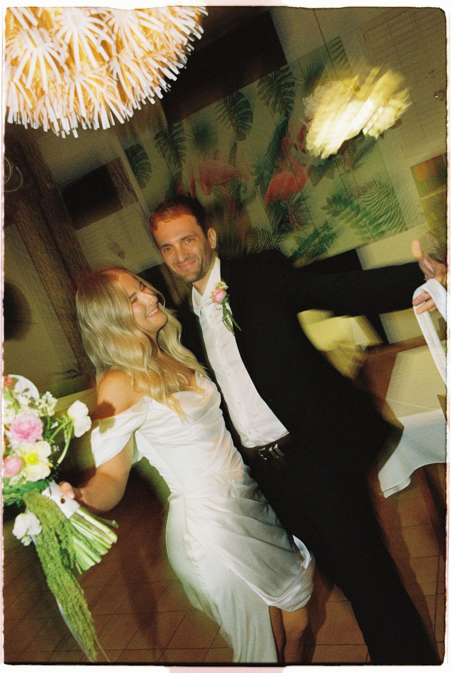 Smiling bride in white dress holding bouquet dancing with groom in black suit at wedding reception.