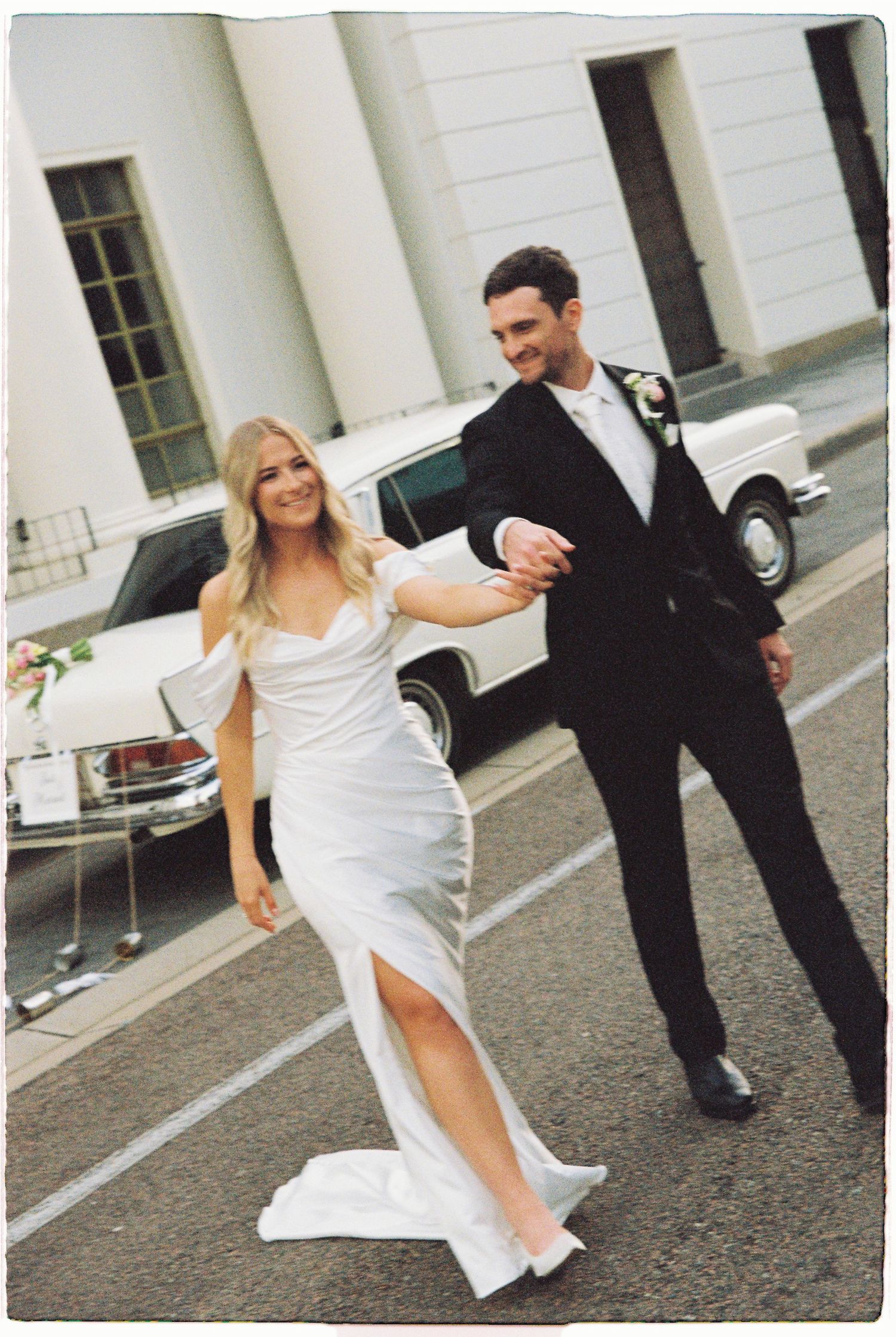 Bride in white slit gown and groom in black tuxedo dancing together beside a vintage white Mercedes wedding car.