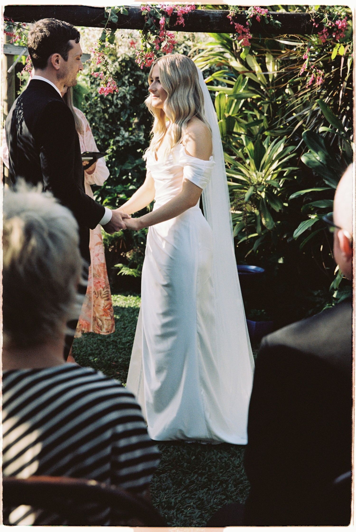 Smiling bride in off-shoulder white gown holding grooms hands during outdoor garden ceremony.