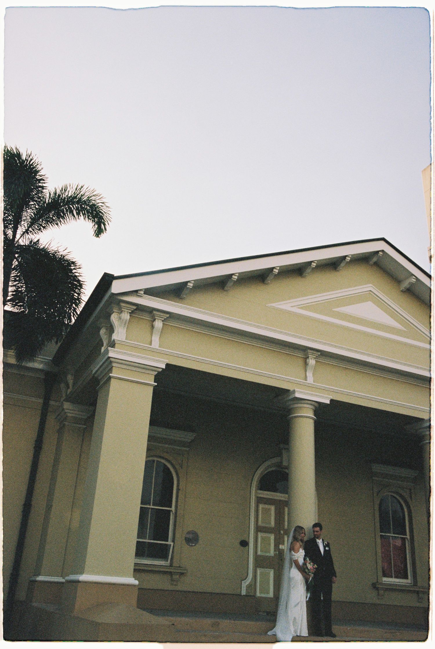 Bride and groom posing outside a yellow colonial building with white columns and a palm tree at golden hour.