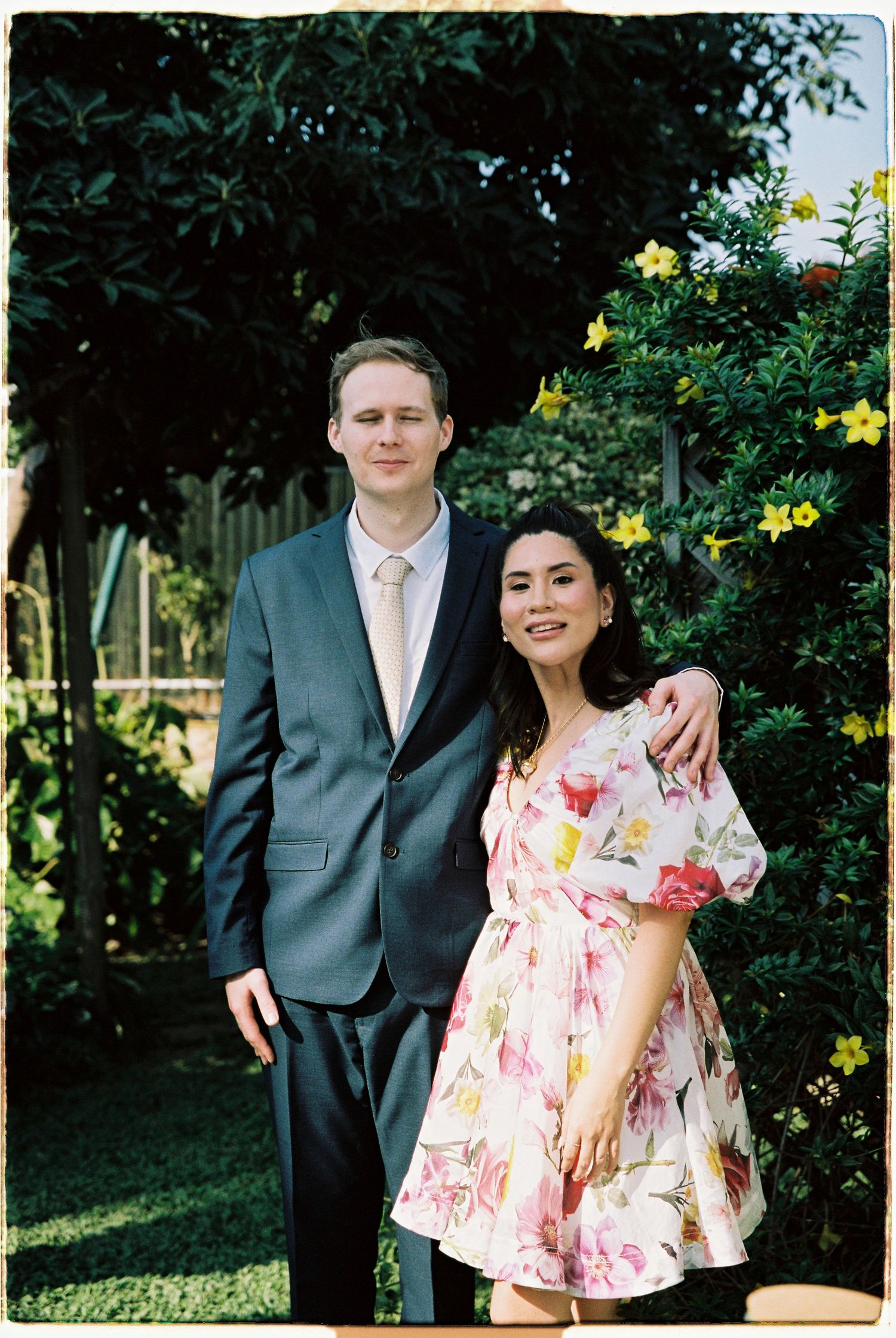 A couple poses together in a sunny garden, man in grey suit, woman in floral dress.