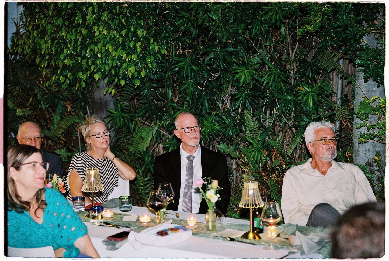 Adults seated at an elegant outdoor dinner table with candles and wine glasses, surrounded by lush green foliage.