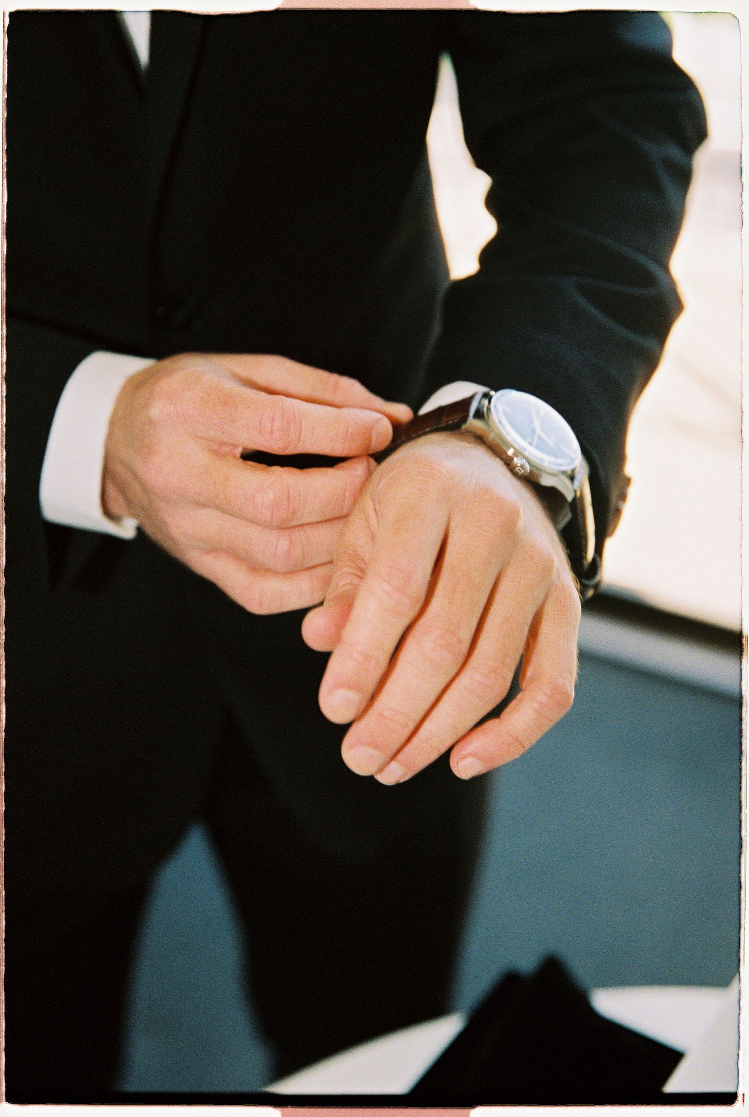 Close-up of groom in black suit adjusting leather wristwatch on his wrist before wedding.