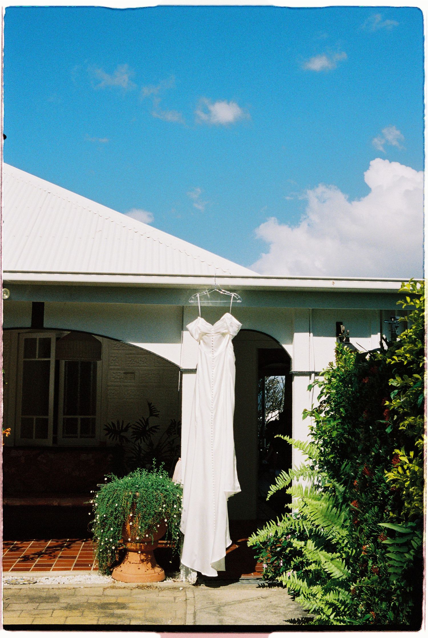 White wedding dress hanging on hanger outside a charming white house under blue sky.