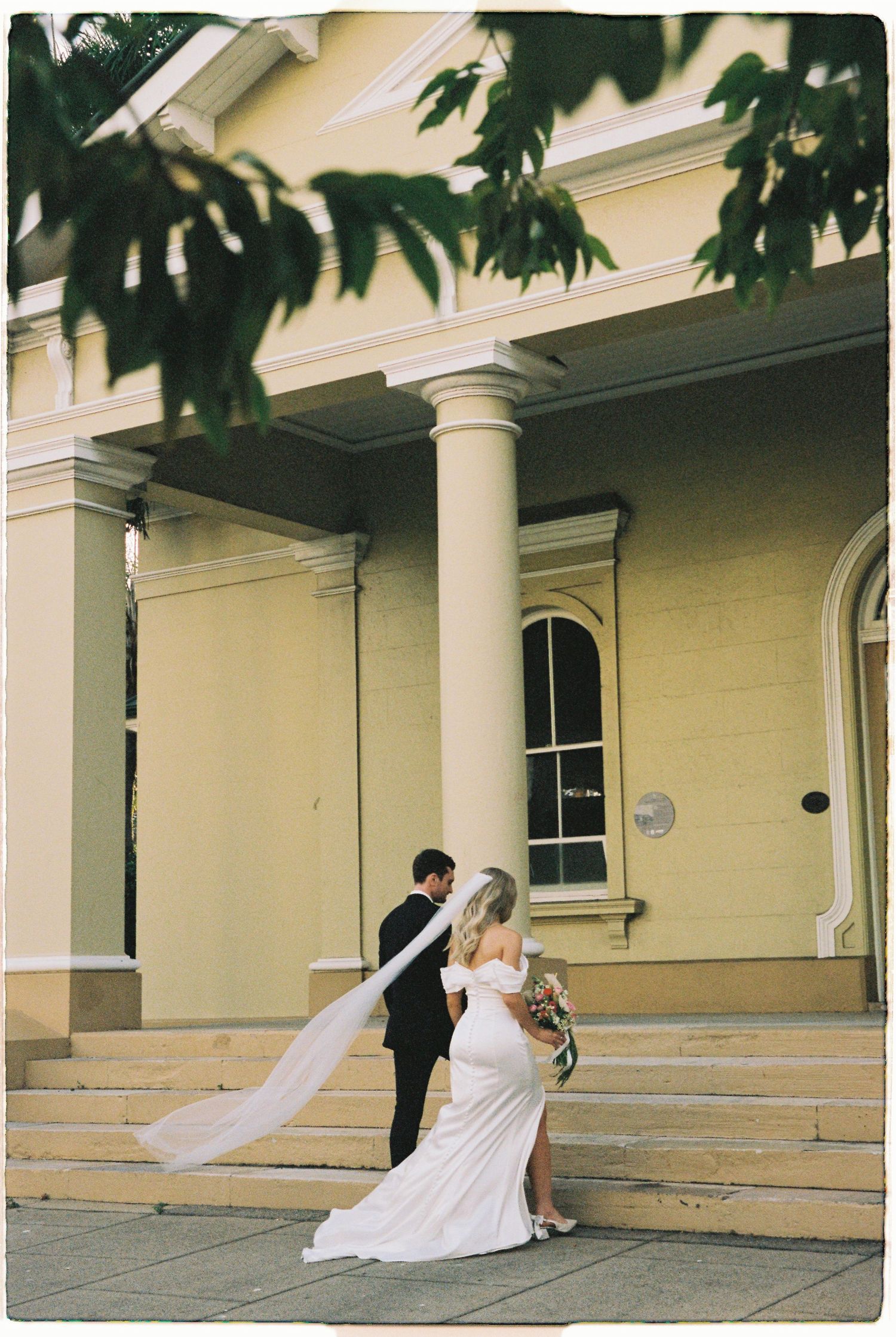 Bride and groom embracing on steps of yellow colonial building with white columns, veil flowing.