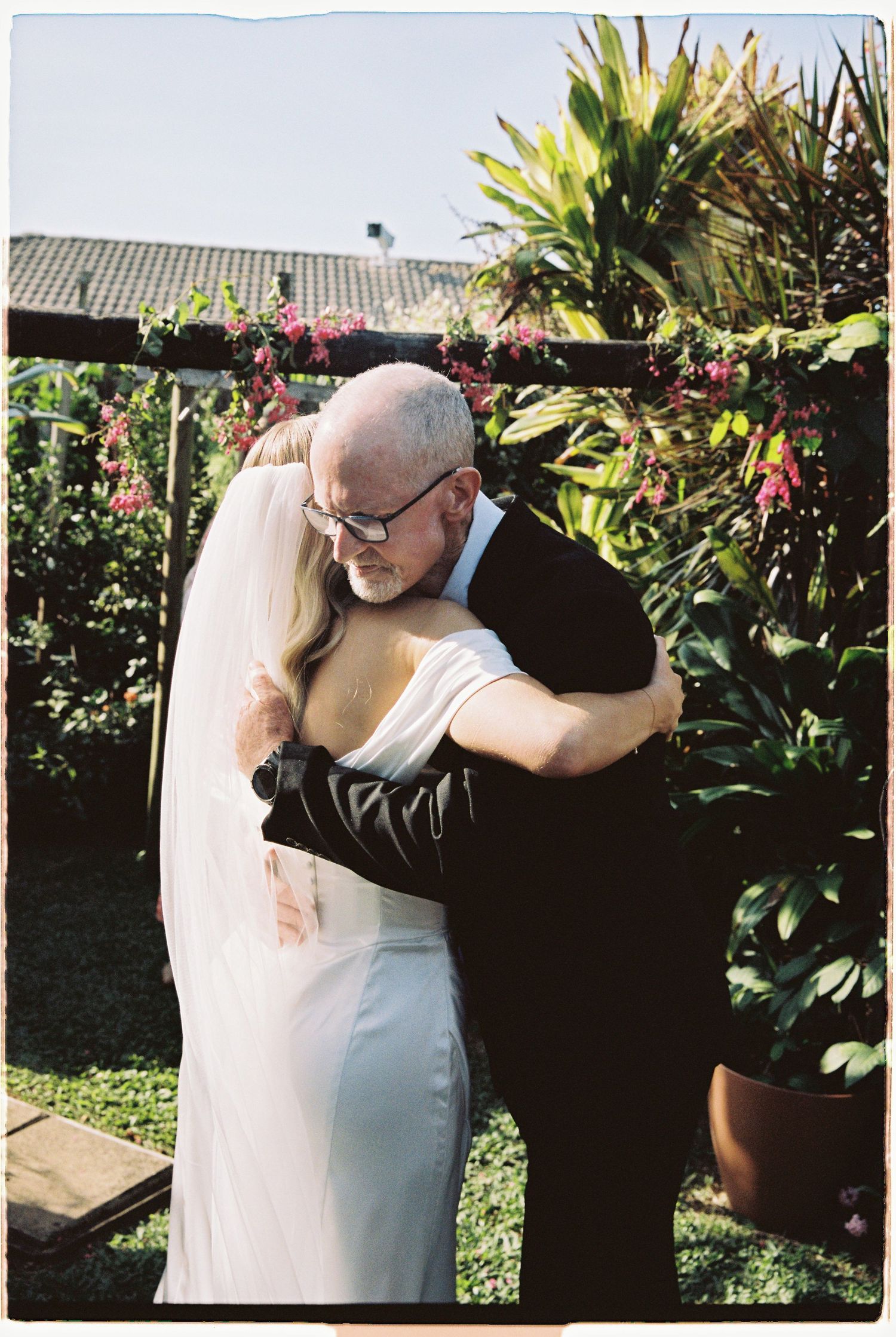 Bride and older man share emotional hug in garden wedding setting with tropical plants.