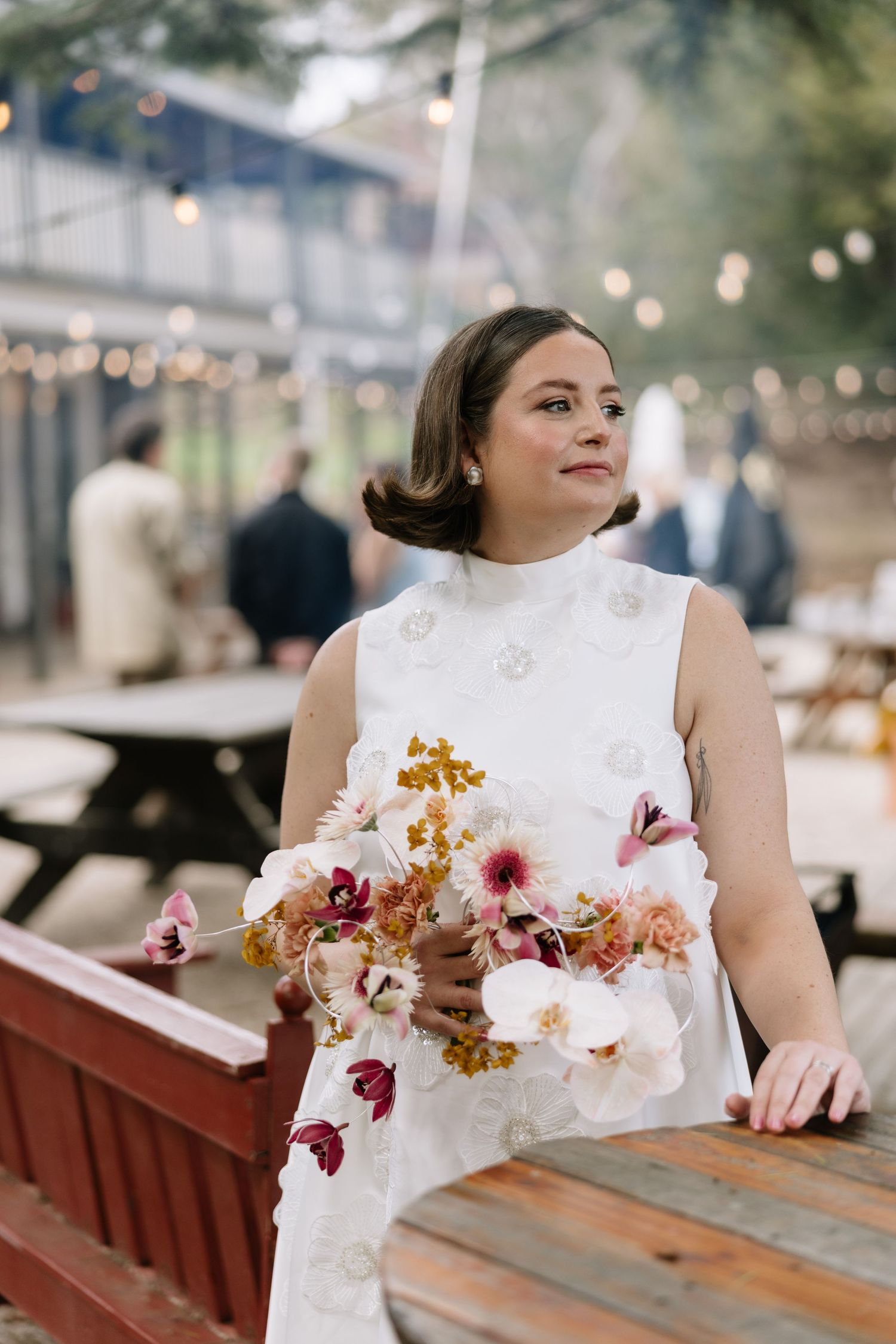Bride in white floral dress holding colorful bouquet of orchids and wildflowers at outdoor wedding venue.