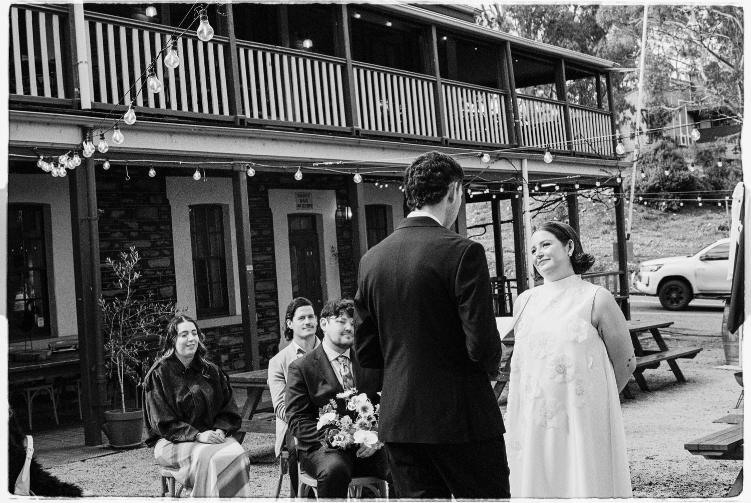 Black and white outdoor wedding ceremony with bride and groom exchanging vows before seated guests near a rustic two-story venue.