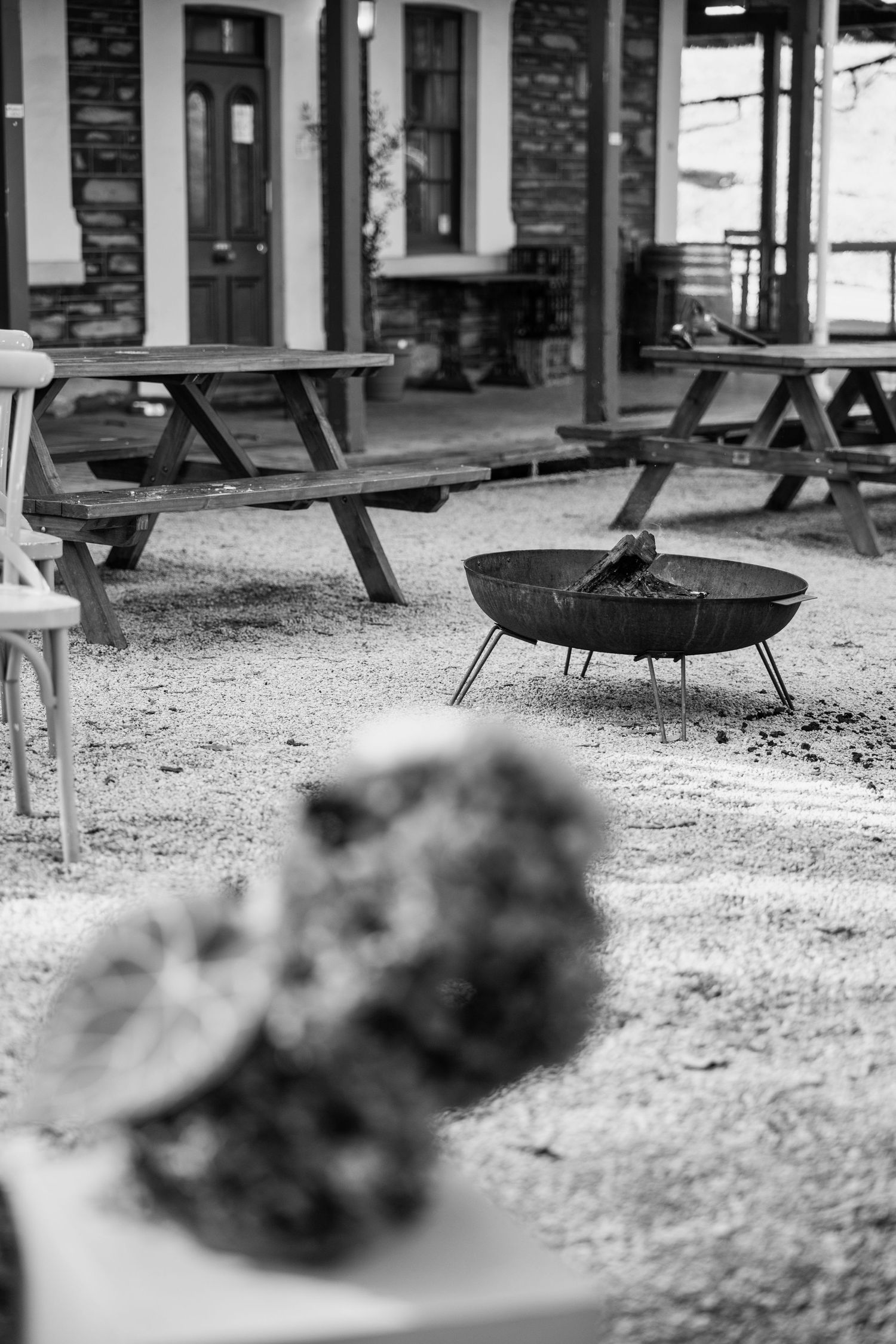 Black and white photo of outdoor patio with picnic tables, fire pit, and blurred floral arrangement in foreground.