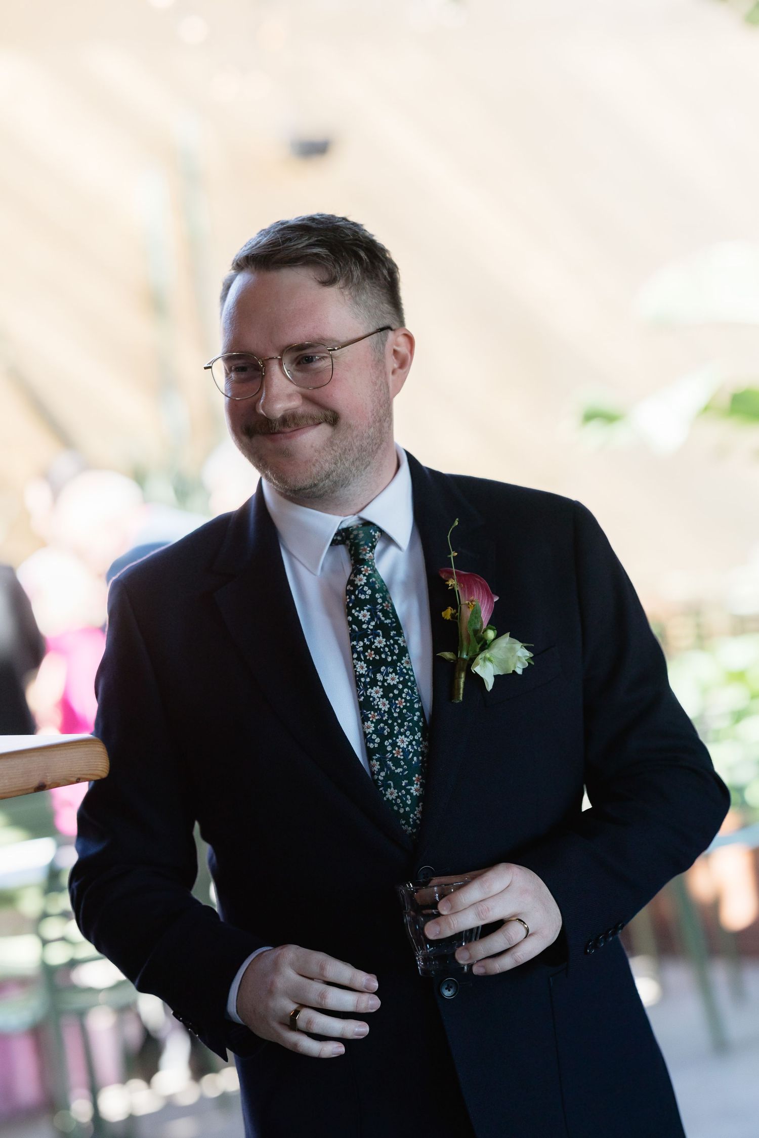 Smiling groom in navy suit with floral tie and pink boutonniere at wedding ceremony.