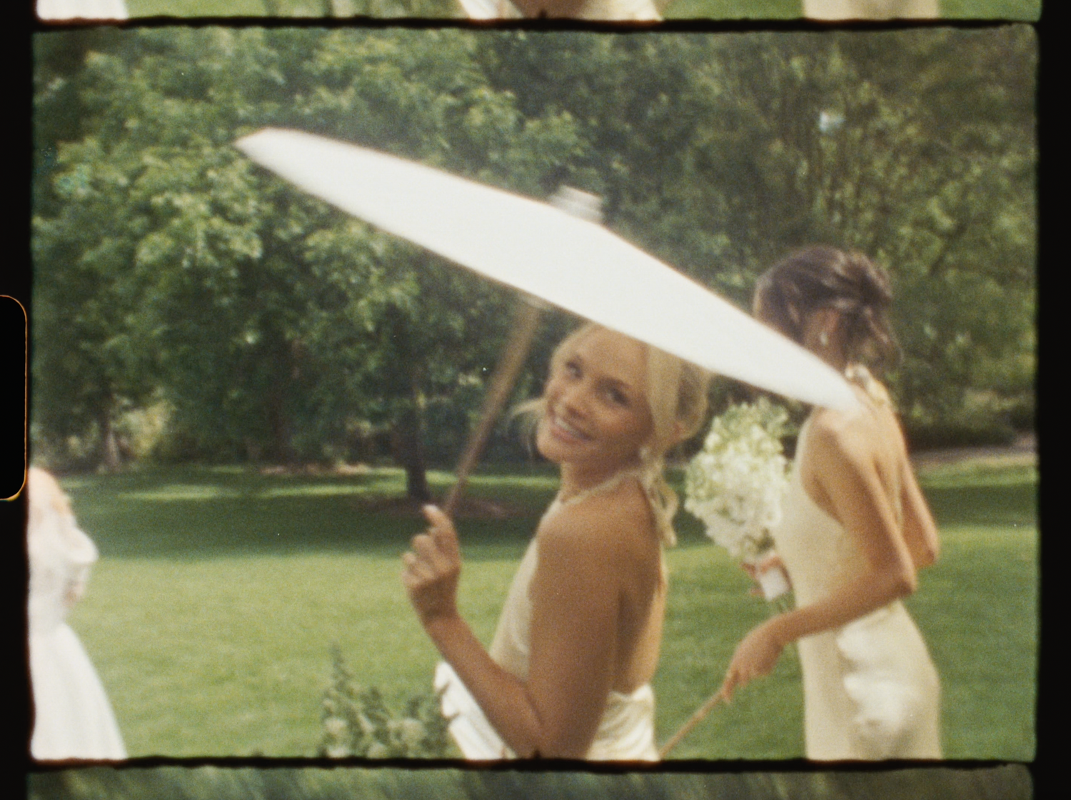Smiling bridesmaid in white dress holding a white parasol umbrella outdoors at a wedding ceremony.