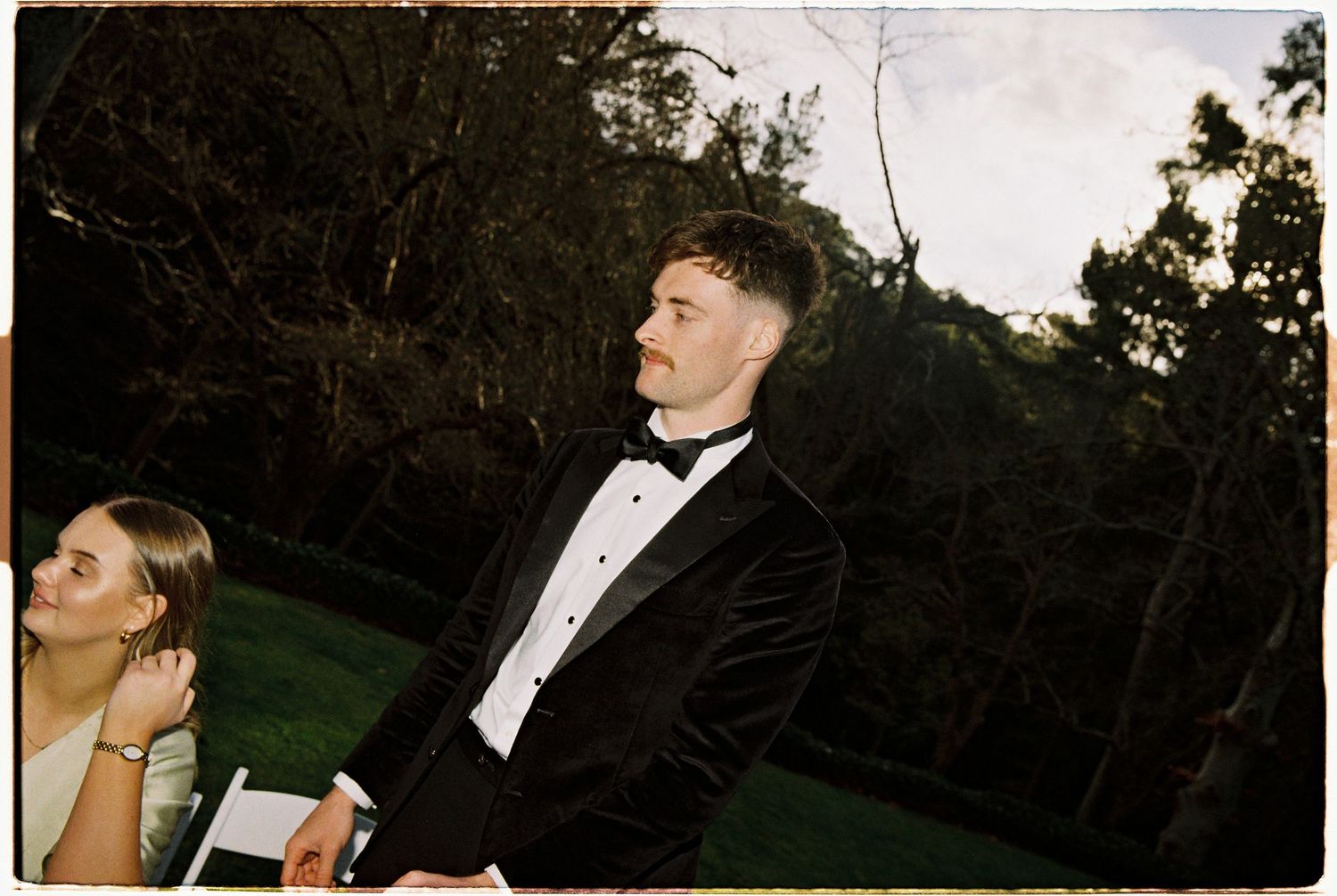 Man in black tuxedo with bow tie standing outdoors at evening wedding reception among lush green trees.