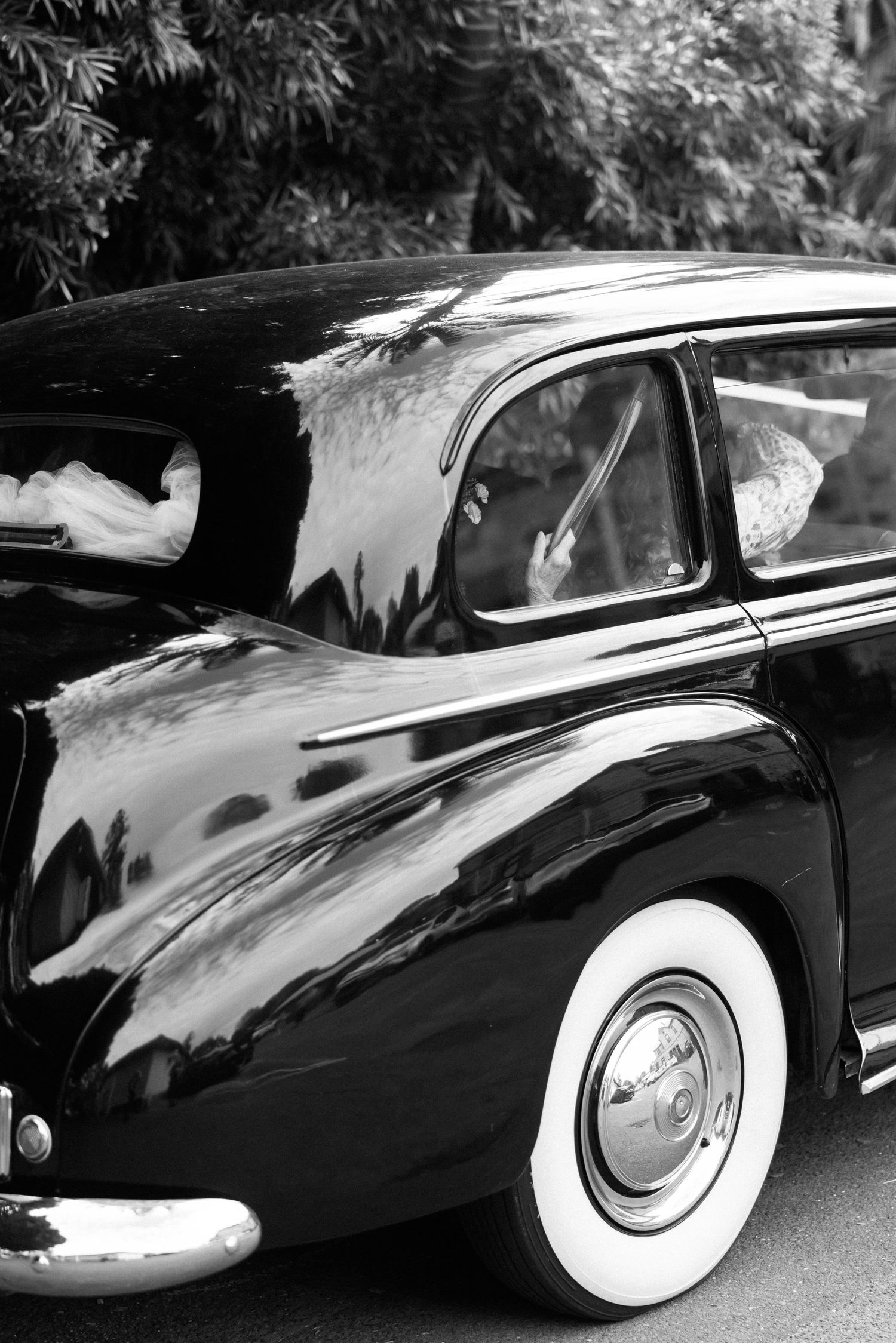Black and white photo of a classic vintage car with white wall tires and shiny chrome details parked outdoors.