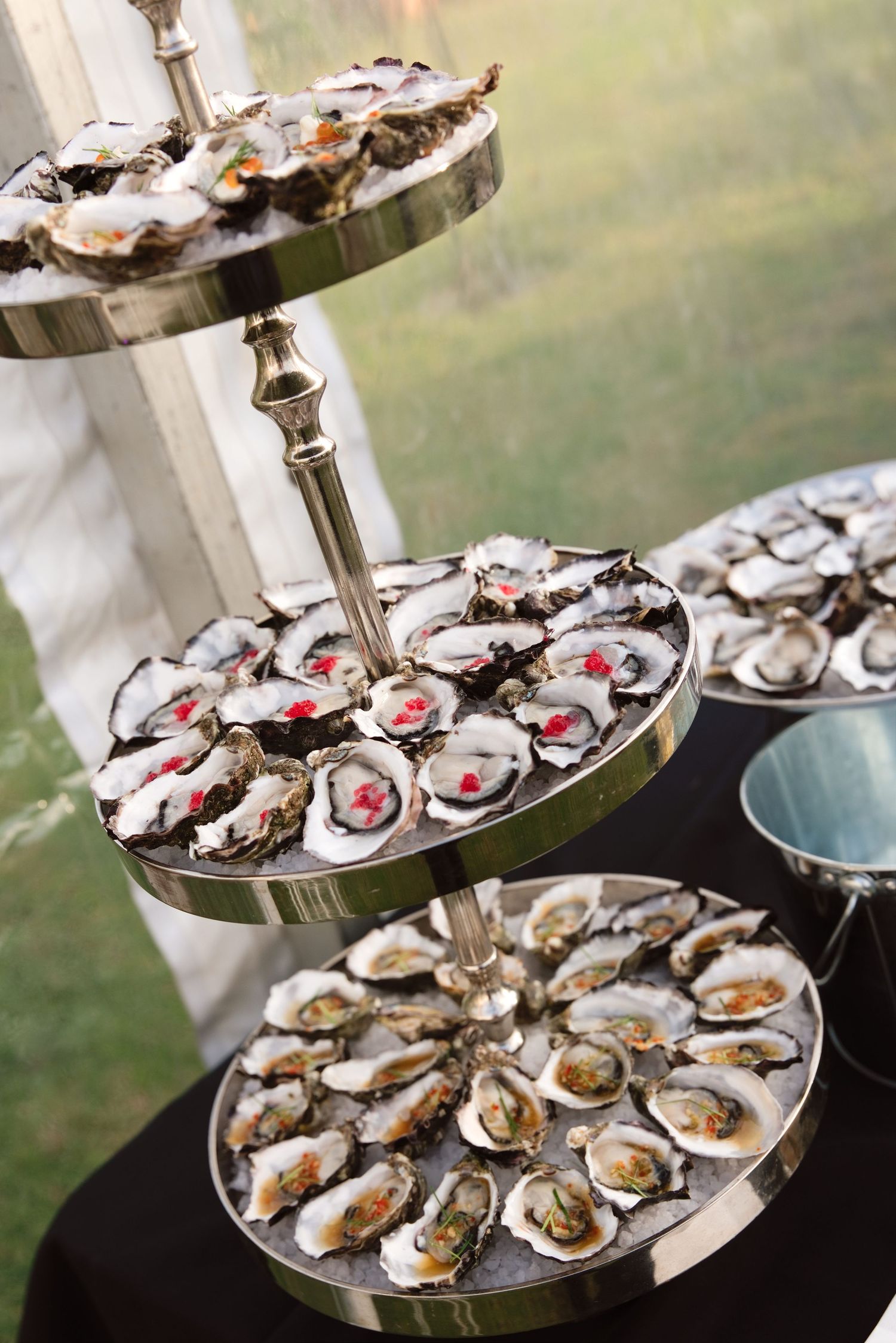 Elegant three-tier silver stand displaying fresh oysters on ice at a catered event or wedding reception.