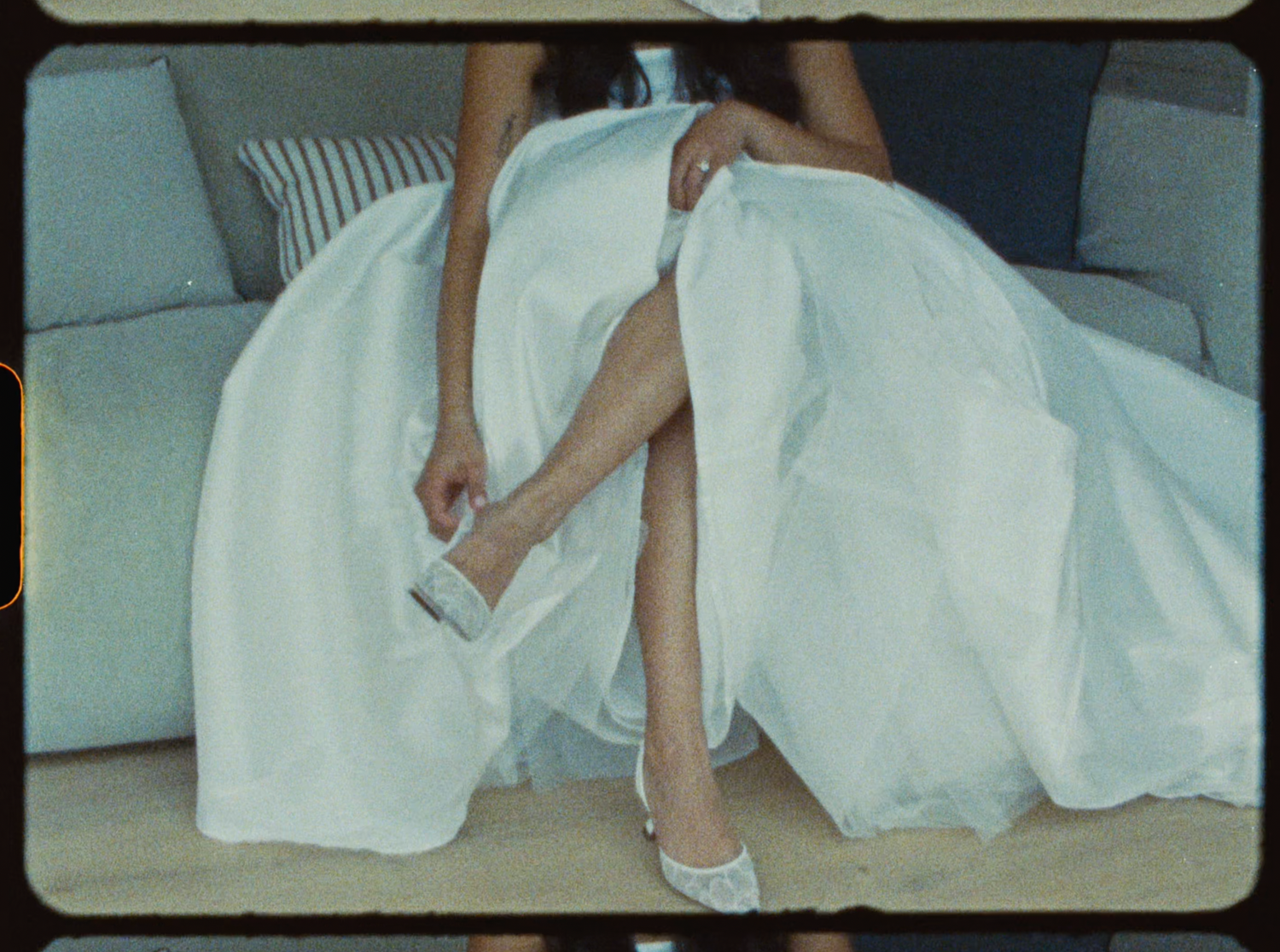 Bride in voluminous white wedding gown adjusting white heels while seated on a sofa.