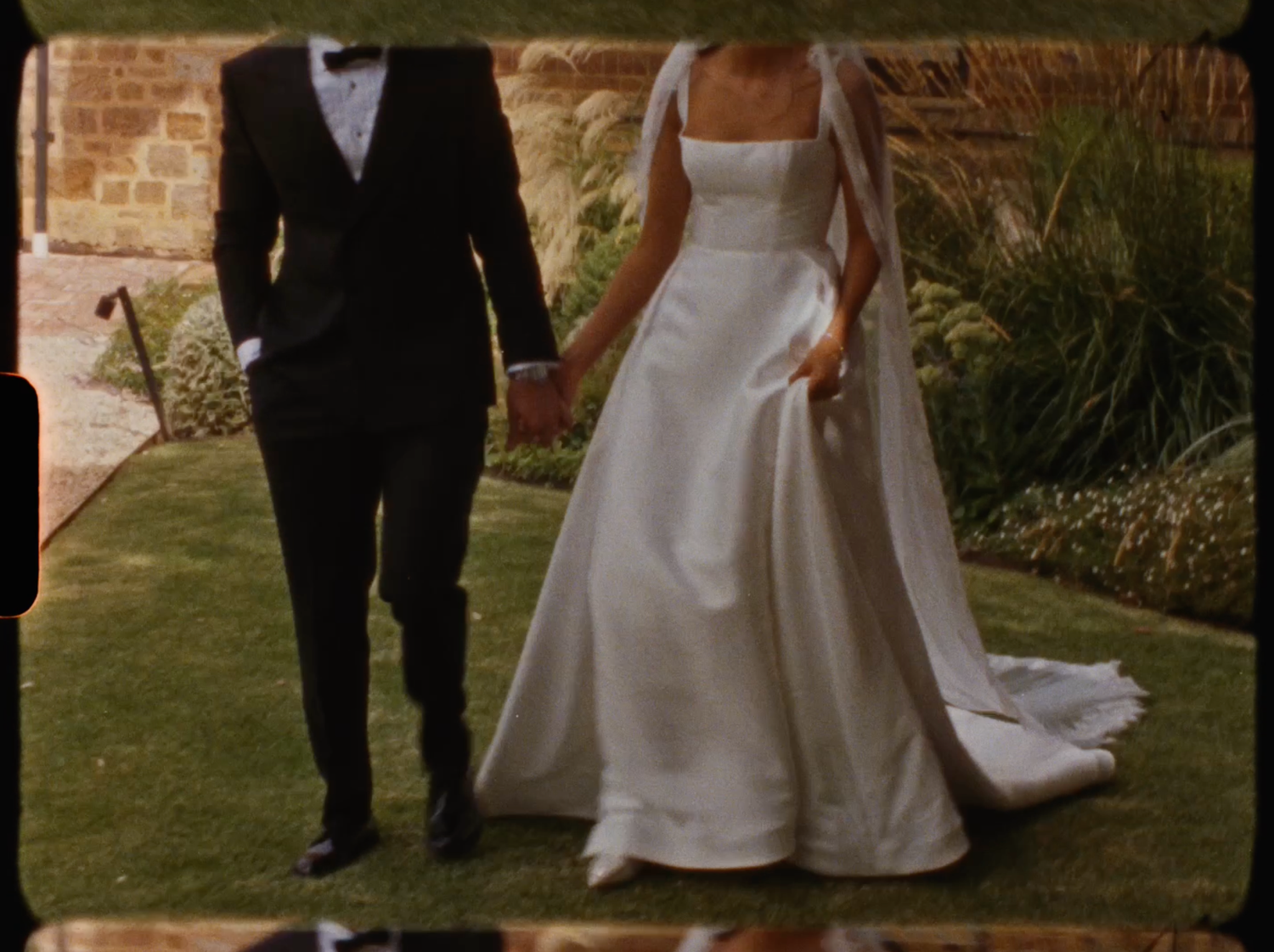 Bride in elegant white satin ball gown holding hands with groom in black tuxedo outdoors.