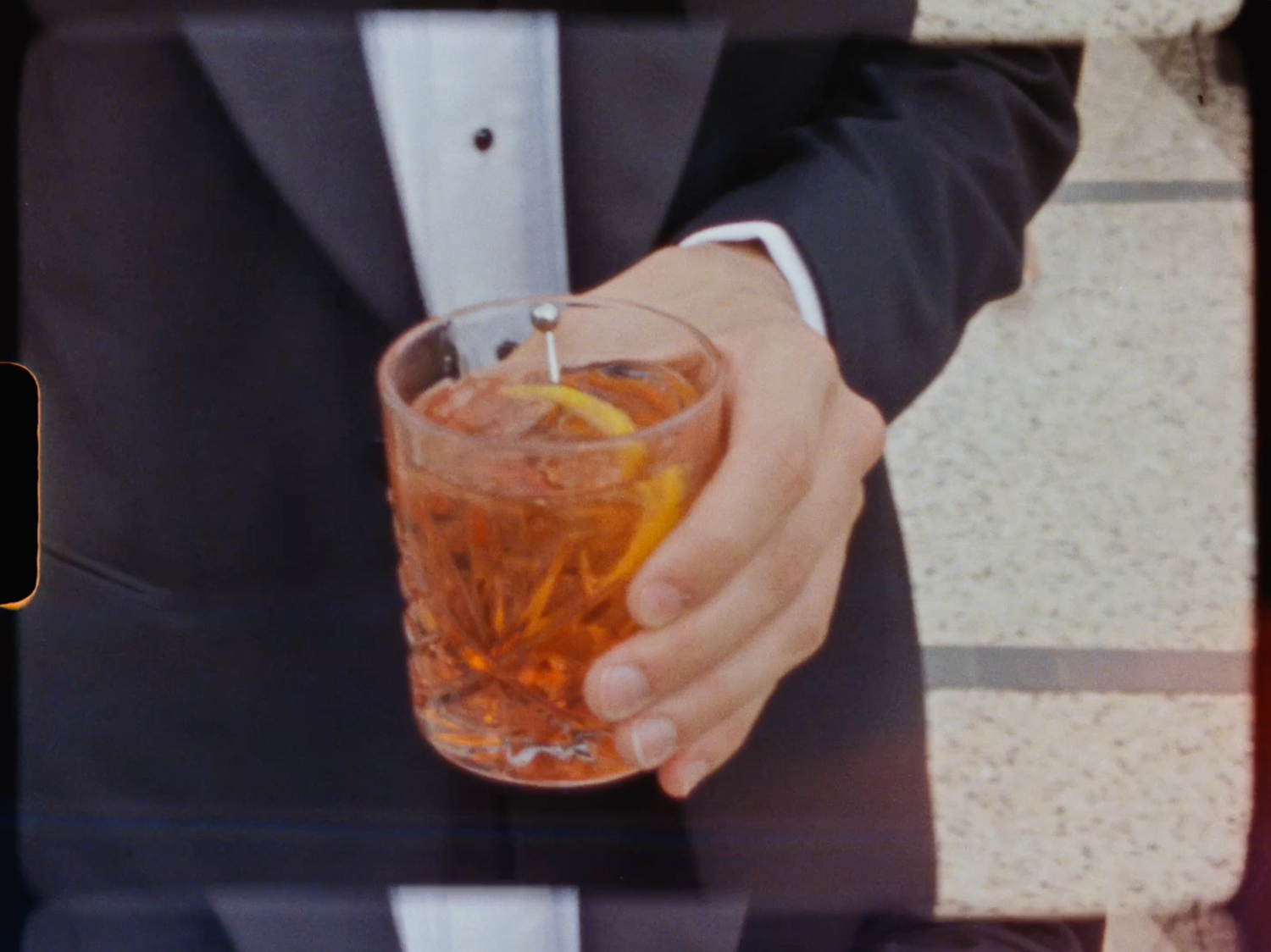 Groom in tuxedo holds a crystal glass with an amber cocktail garnished with orange peel at a wedding.