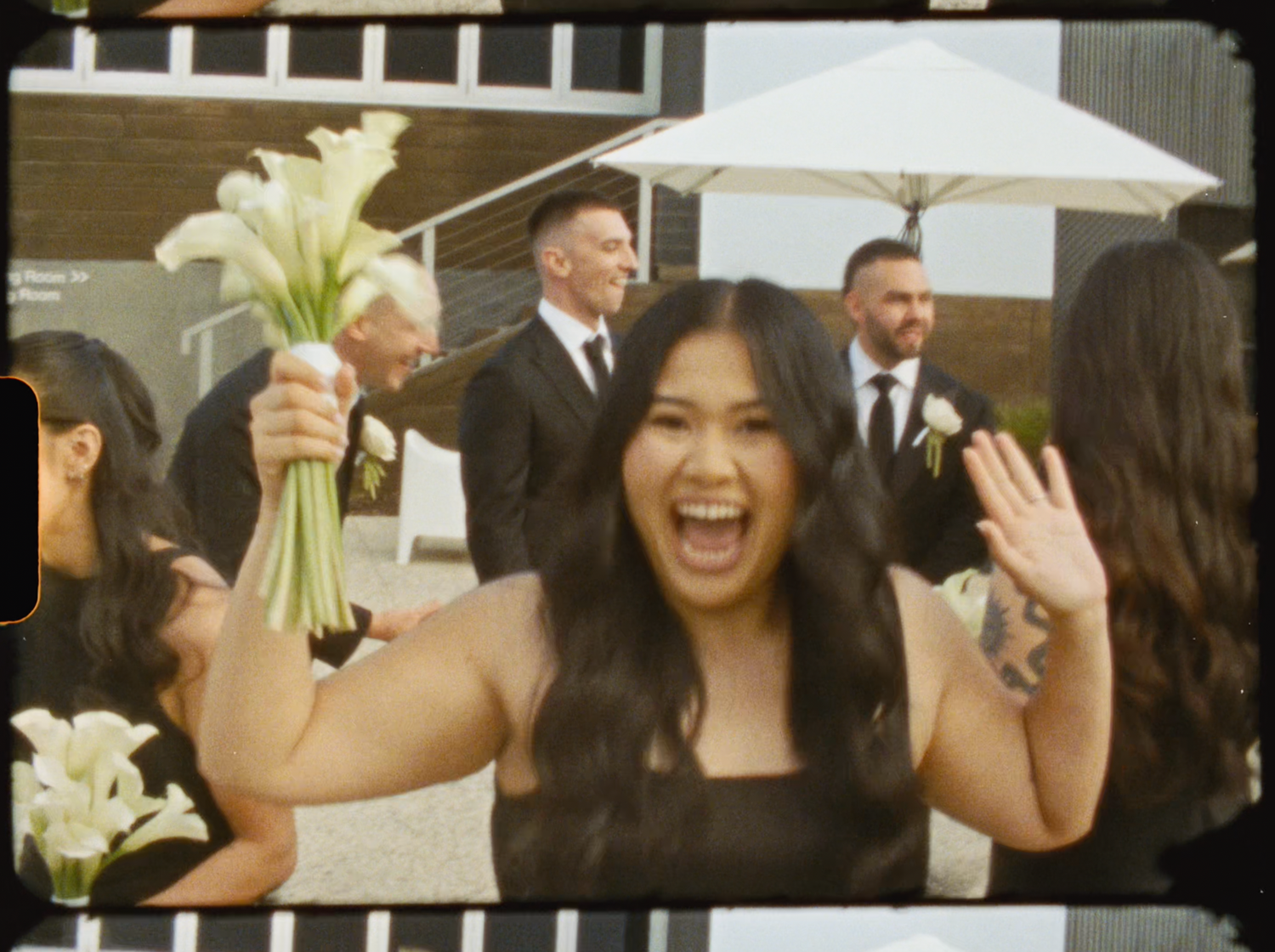 Excited bridesmaid in black dress laughs and waves while holding white calla lily bouquet at wedding.