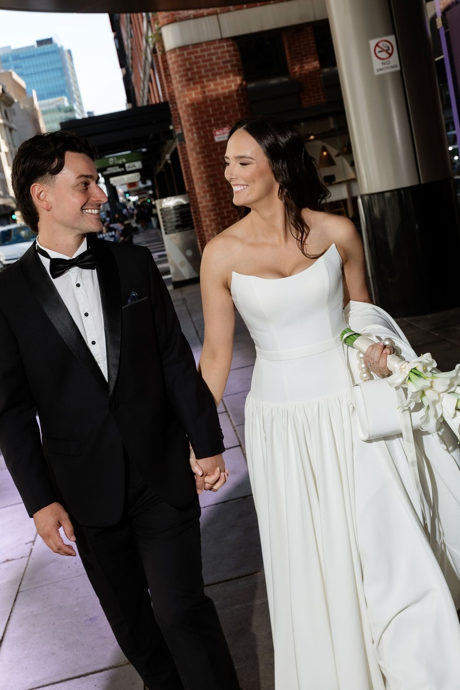 Bride and groom holding hands and smiling while walking on a city street on their wedding day.