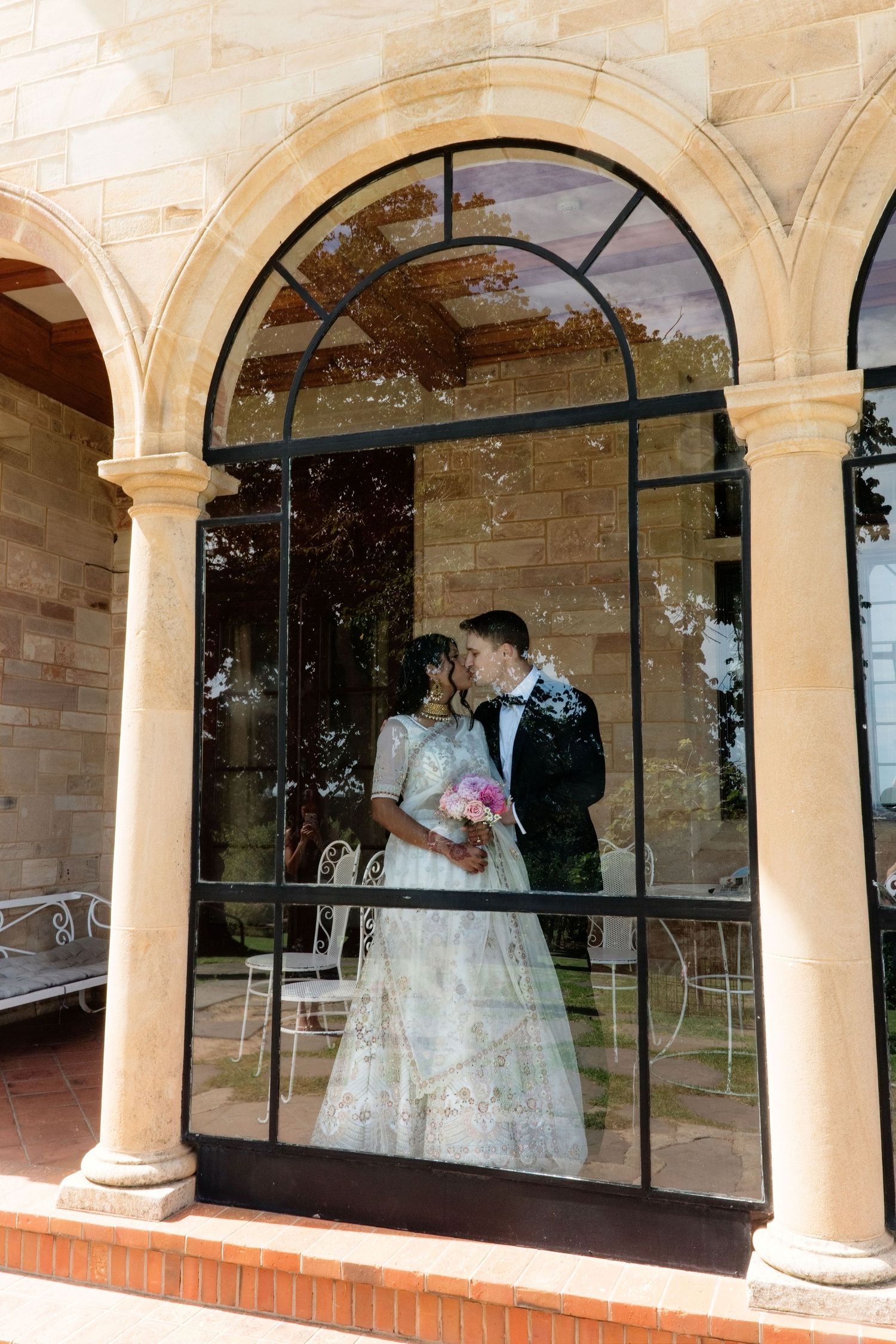 Bride and groom sharing a kiss reflected in large arched glass window of elegant sandstone wedding venue.