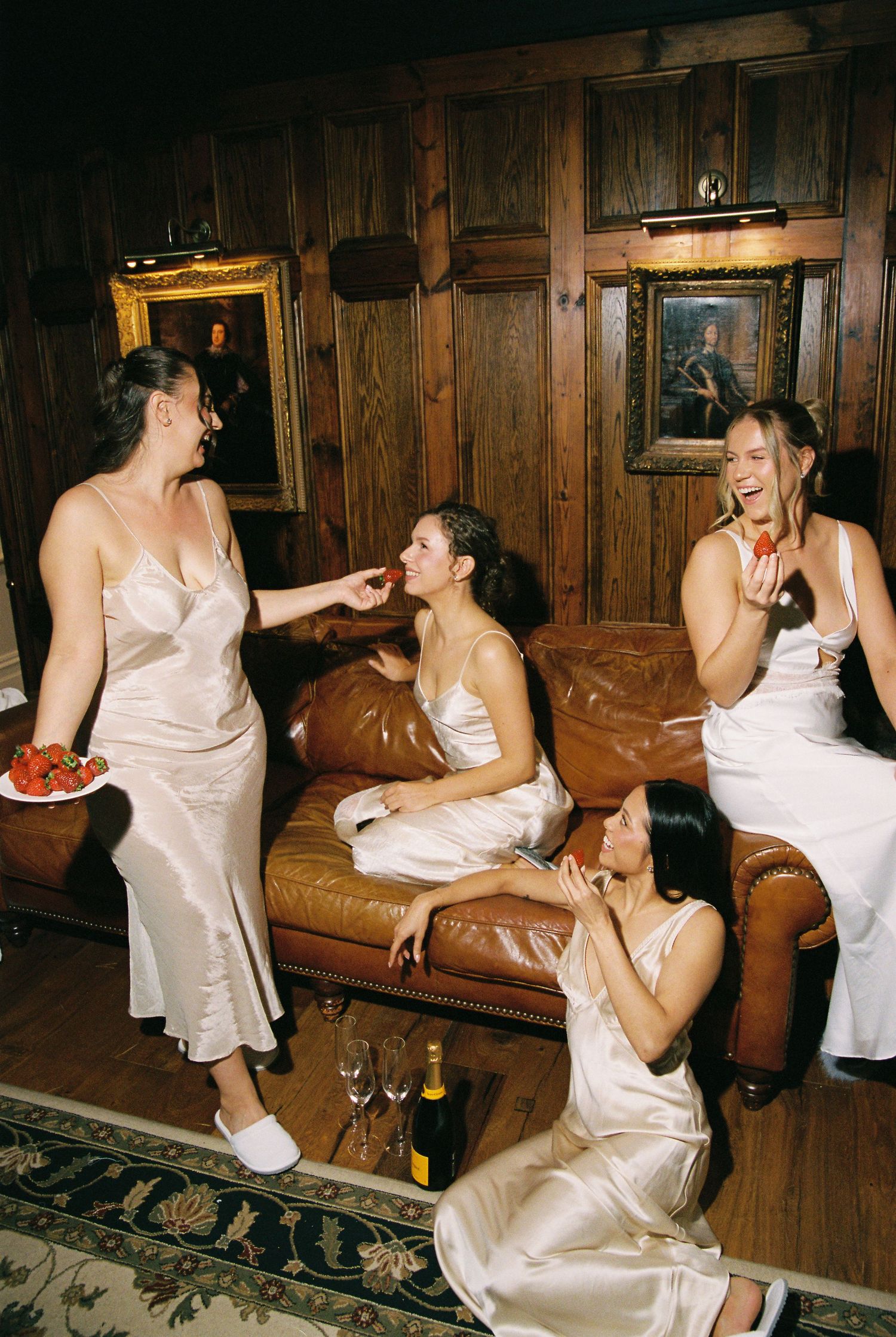 Bridesmaids in silk slip dresses sharing strawberries and champagne on a leather sofa.