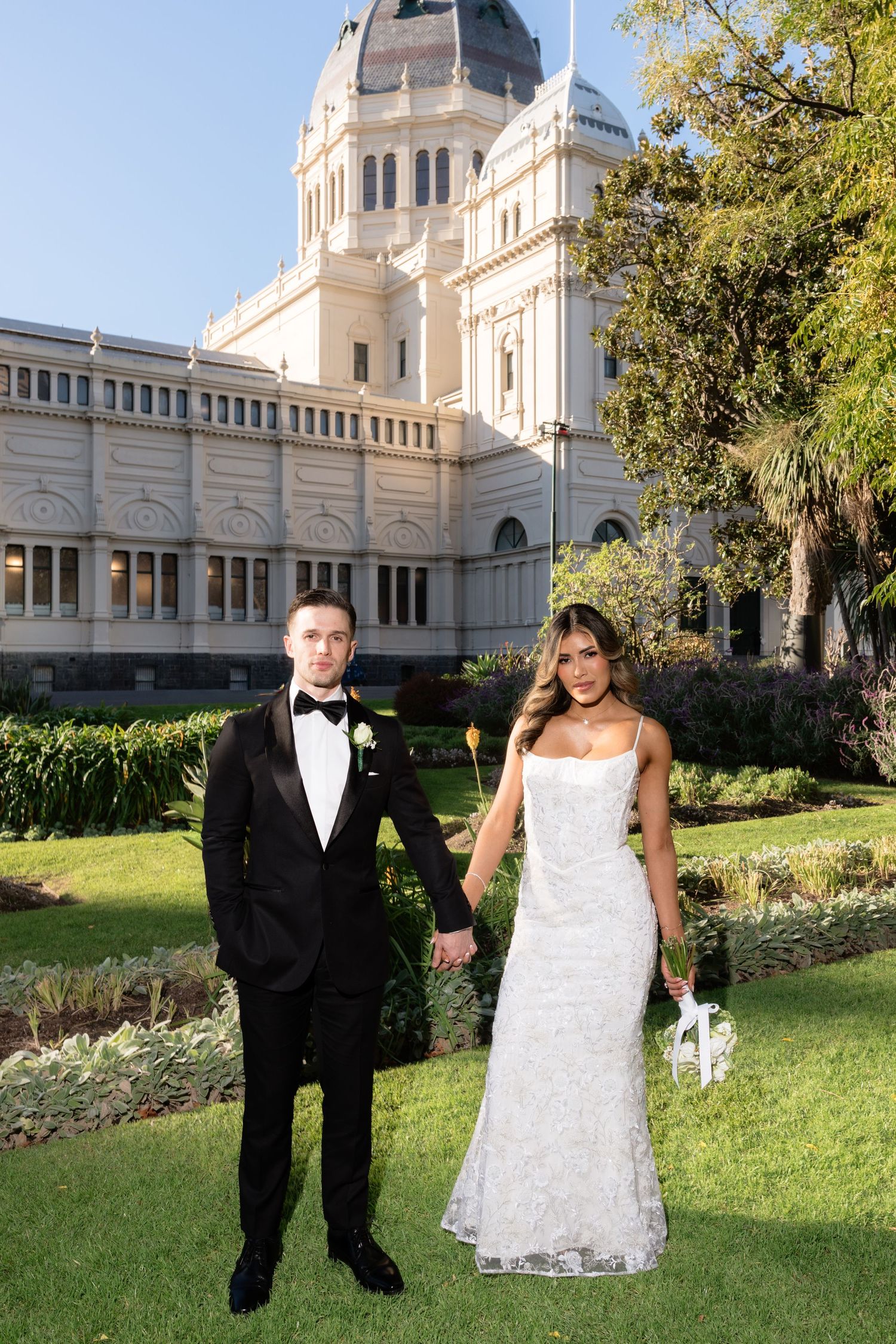 Wedding couple holding hands in formal attire before the Royal Exhibition Building Melbourne gardens.