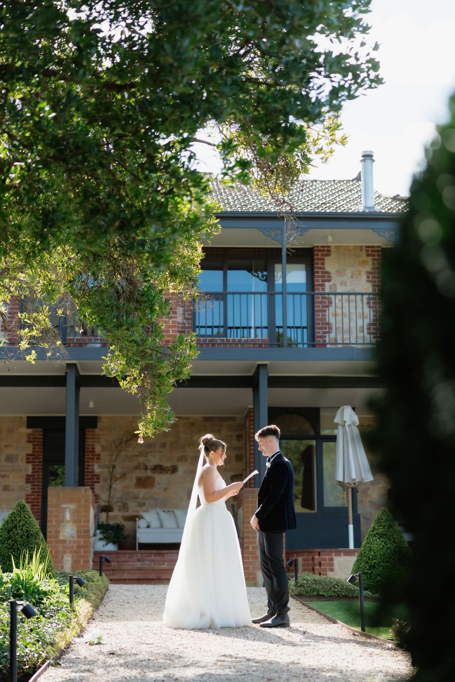 Bride and groom exchange vows outdoors in front of a rustic brick venue surrounded by lush green trees.