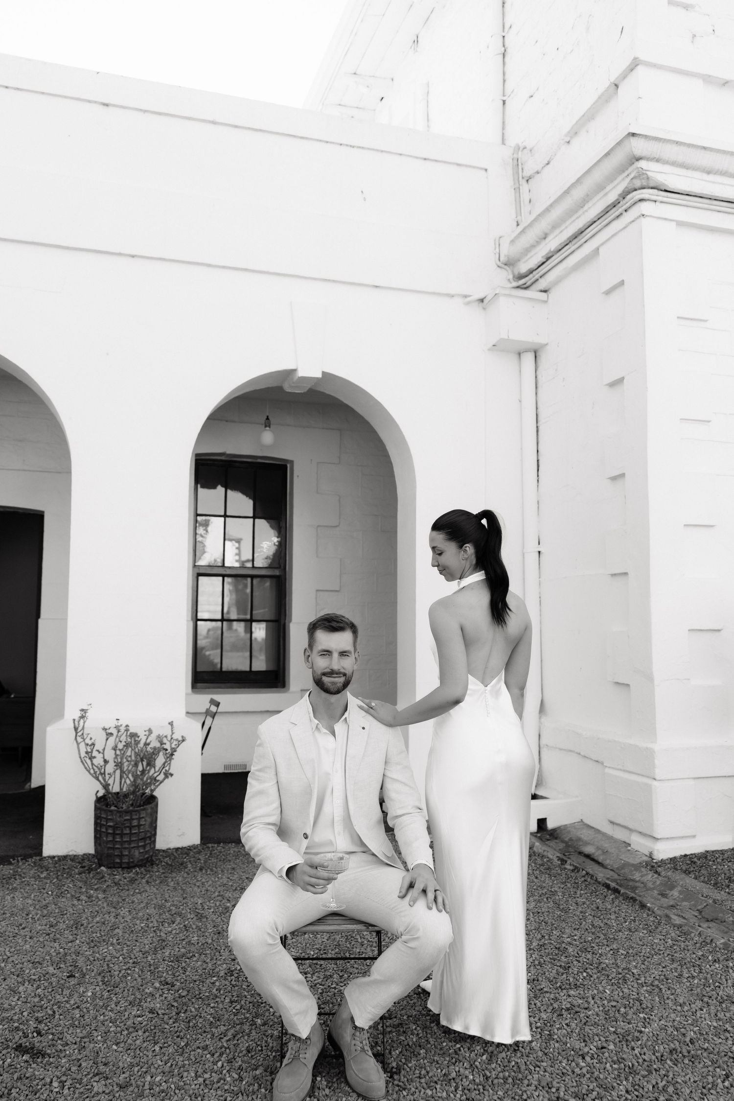 Black and white photo of a groom sitting outdoors while bride in backless gown stands beside him at elegant venue.