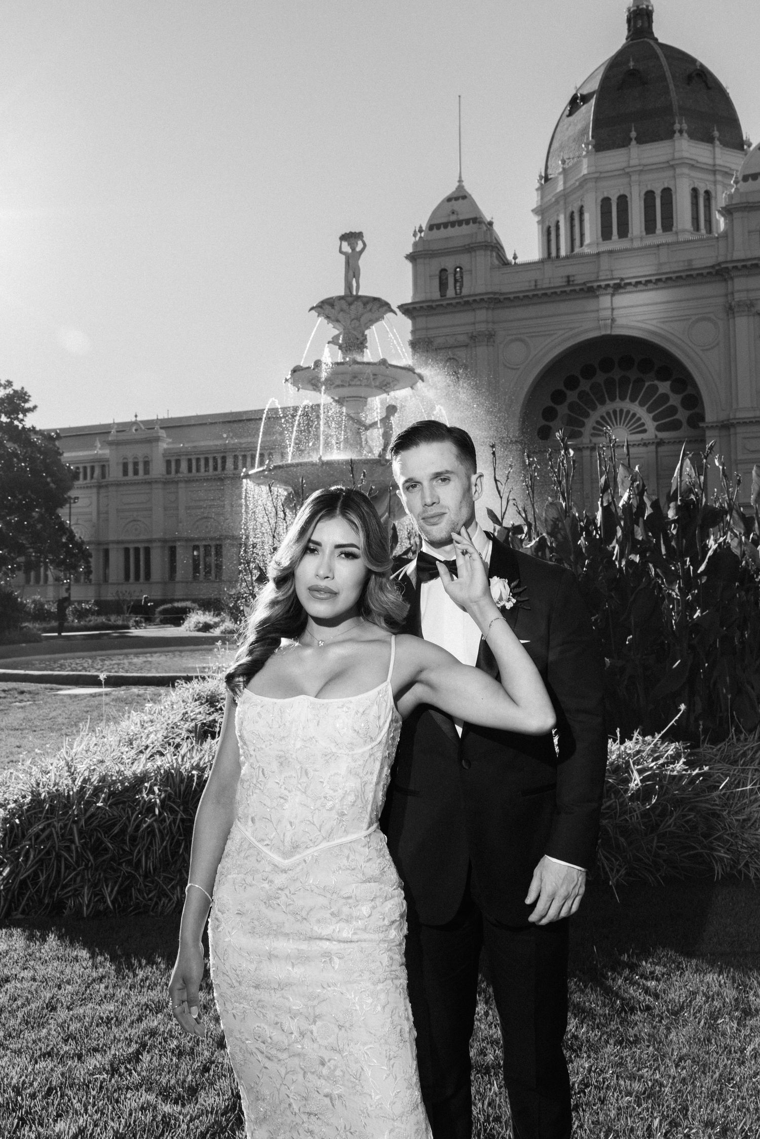 Black and white wedding photo of bride and groom posing before ornate fountain and domed building.