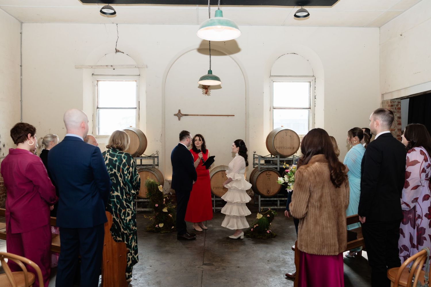 Wedding ceremony in a rustic winery with wooden barrels, guests standing as couple exchanges vows under pendant lights.