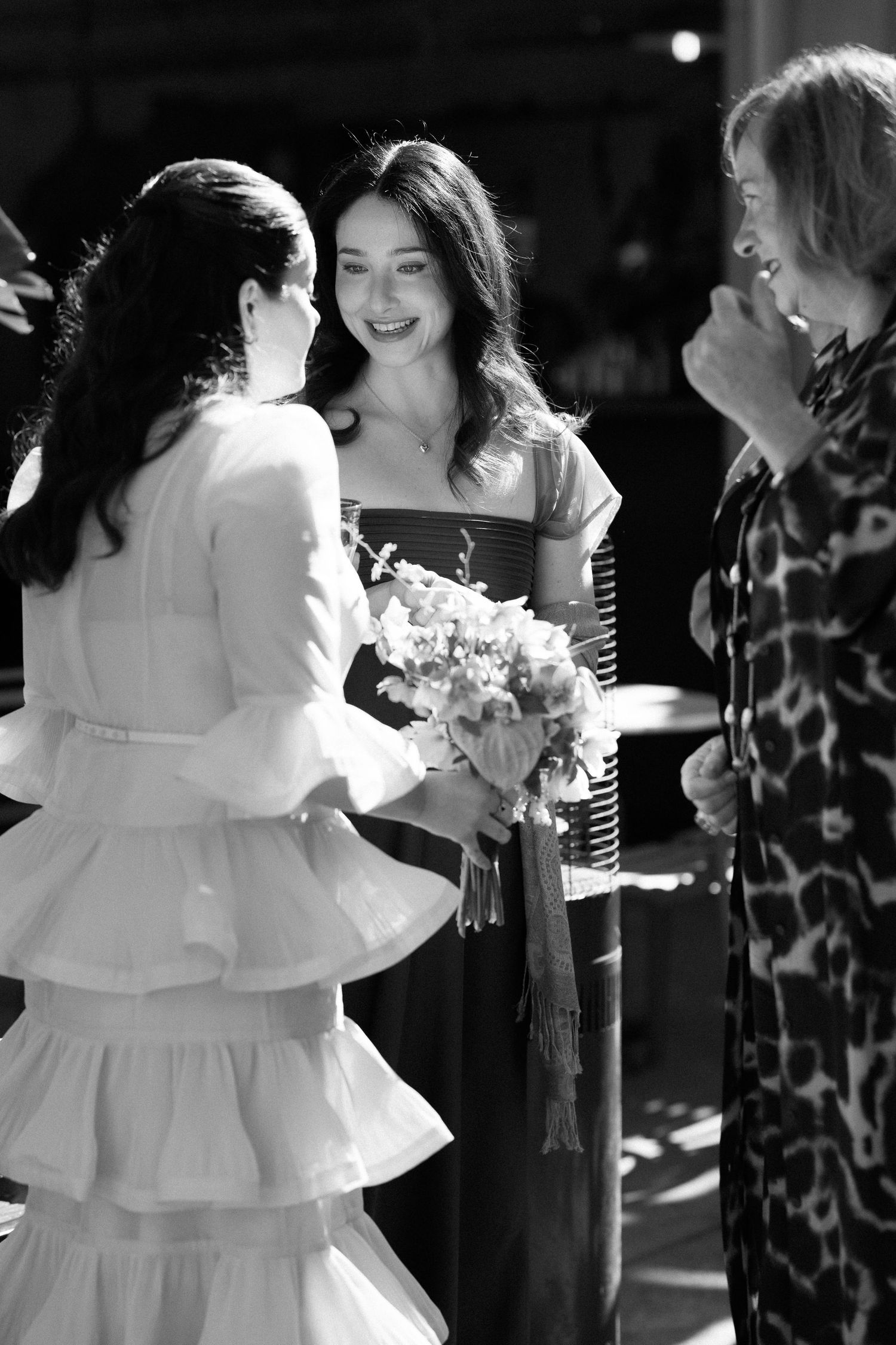 Black and white photo of bride in ruffled dress greeting guests while holding wedding bouquet.