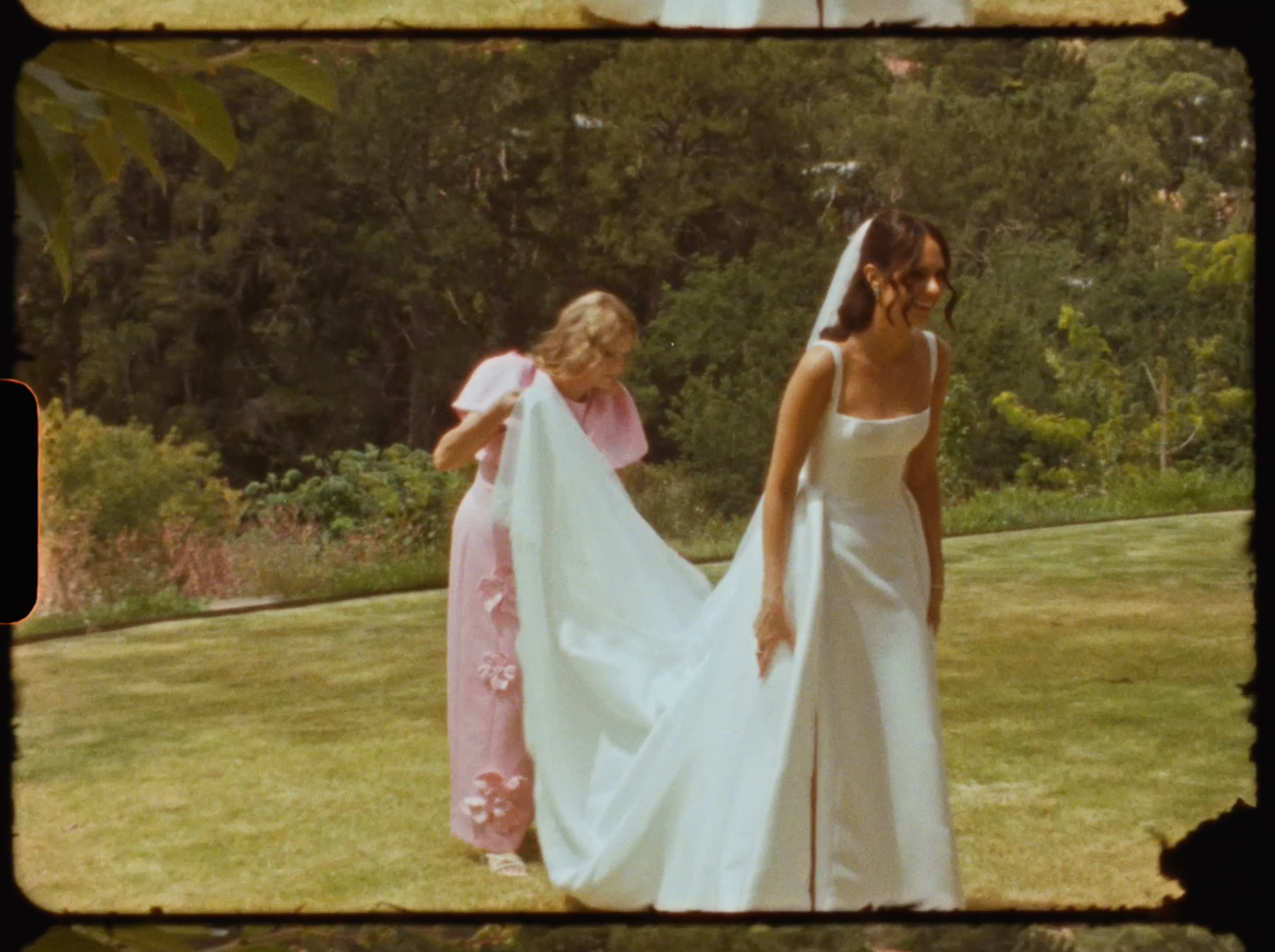 Bridesmaid in pink dress arranges the train of a bride's white gown on green lawn.