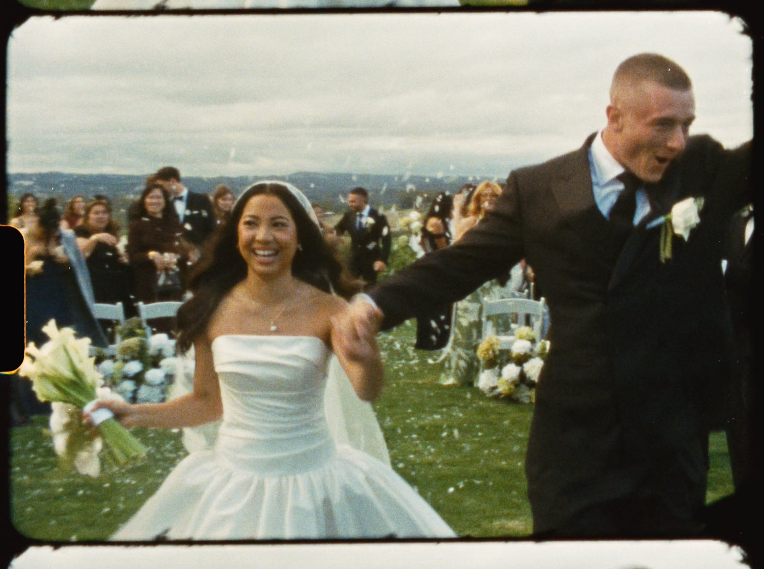 Joyful bride in white strapless gown holding bouquet walking with groom after outdoor wedding ceremony.